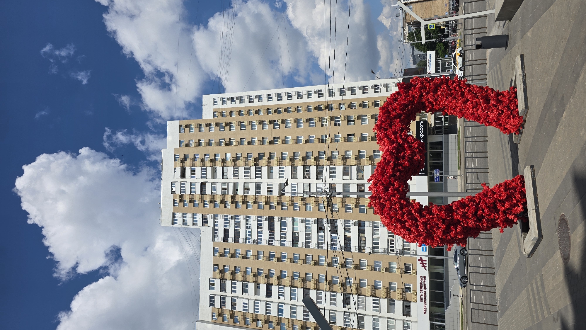 An outdoor, eye-level, vertical, medium shot shows a large, bright red heart-shaped sculpture made of artificial flowers in the foreground. The sculpture is centrally placed and takes up a significant portion of the lower half of the frame. Behind the sculpture, a tall, beige and white apartment building dominates the background. The building has many windows and balconies. A bright blue sky with fluffy white clouds is visible above the building. In the middle ground, there is a street with cars and a sidewalk. The scene appears to be in a city, likely Moscow, Russia, given the architectural style of the building and the presence of Cyrillic text on signs. The overall impression is a sunny day in an urban environment, with the vibrant heart sculpture adding a decorative element.