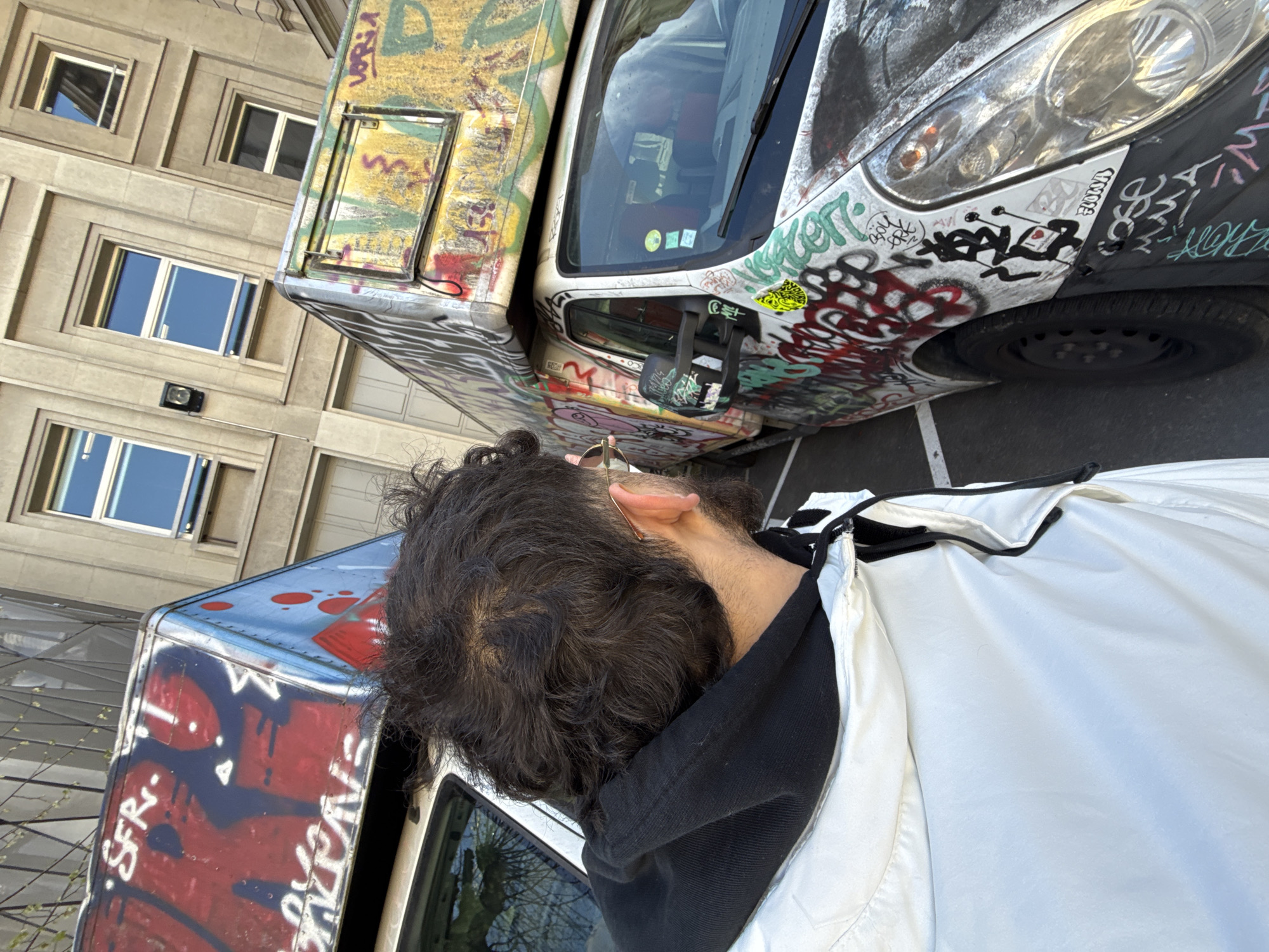 The image depicts an urban outdoor scene in Strasbourg, France. The foreground shows the rear and right profile of an individual with dark, curly hair exhibiting partial balding, a beard, and thin, gold-framed glasses. This person wears a white jacket over a black hooded sweatshirt, with their head turned towards the right.
Two white commercial vans are parked on asphalt pavement in the middle ground, both extensively covered in multi-colored graffiti tags and designs. The van on the left has a red and blue painted upper section with "SFR" lettered in white. The right van, positioned closer to the viewer, displays graffiti across its front bumper, headlights, and side panels, extending to a raised roof compartment adorned with yellow, green, and red graffiti. Its windshield contains several small stickers and reflections. A white parking space line is visible on the asphalt beneath the front wheel of the right van.
The background features a multi-story building constructed from light-toned stone or concrete, characterized by a uniform grid of recessed rectangular windows with light-colored frames. A black wall-mounted light fixture is visible on the building's facade.