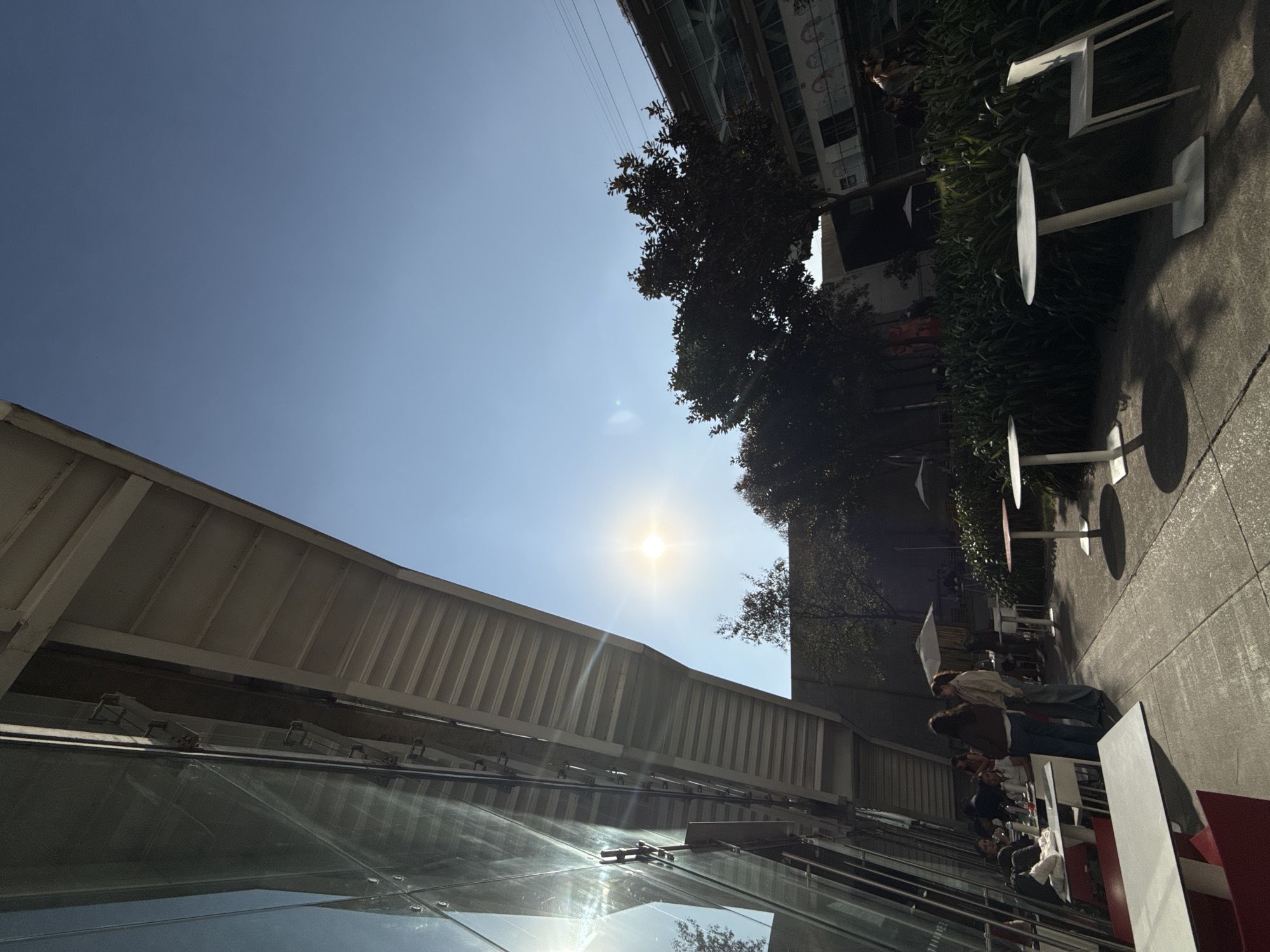 A wide-angle photograph taken on a clear, sunny day depicts an outdoor patio and walkway area alongside a modern architectural structure, identified as likely being within the Santa Fe district of Mexico City, Mexico, consistent with the Tecnológico de Monterrey campus.
The prominent sun is visible in the upper-left quadrant, creating a bright flare on the glass facade of the building to the left and casting distinct shadows across the scene. This building features a steeply angled glass wall reflecting the sunlight, below which a white, ribbed underside of an overhead structure extends horizontally.
The ground is paved with concrete. To the right of the walkway, a dense border of dark green shrubs and trees lines a planter bed. Further in the background, additional trees and a multi-story building with visible windows and upper-level structures are present.
Several white outdoor tables are scattered across the paved area; some are round with central pedestals, others are rectangular. White outdoor chairs accompany these tables, with at least one visible chair standing alone. Two white patio umbrellas are open in the mid-ground, providing shade.
In the foreground-left, a group of individuals is seated at rectangular tables beneath the building's overhang. Visible items on these tables include white napkins and bottles. In the mid-ground, two individuals stand on the walkway engaged in apparent conversation; one wears a dark jacket and blue jeans, facing away from the camera, while the other wears a light-colored top and light jeans, facing the first individual. Additional indistinct figures are present in the background.