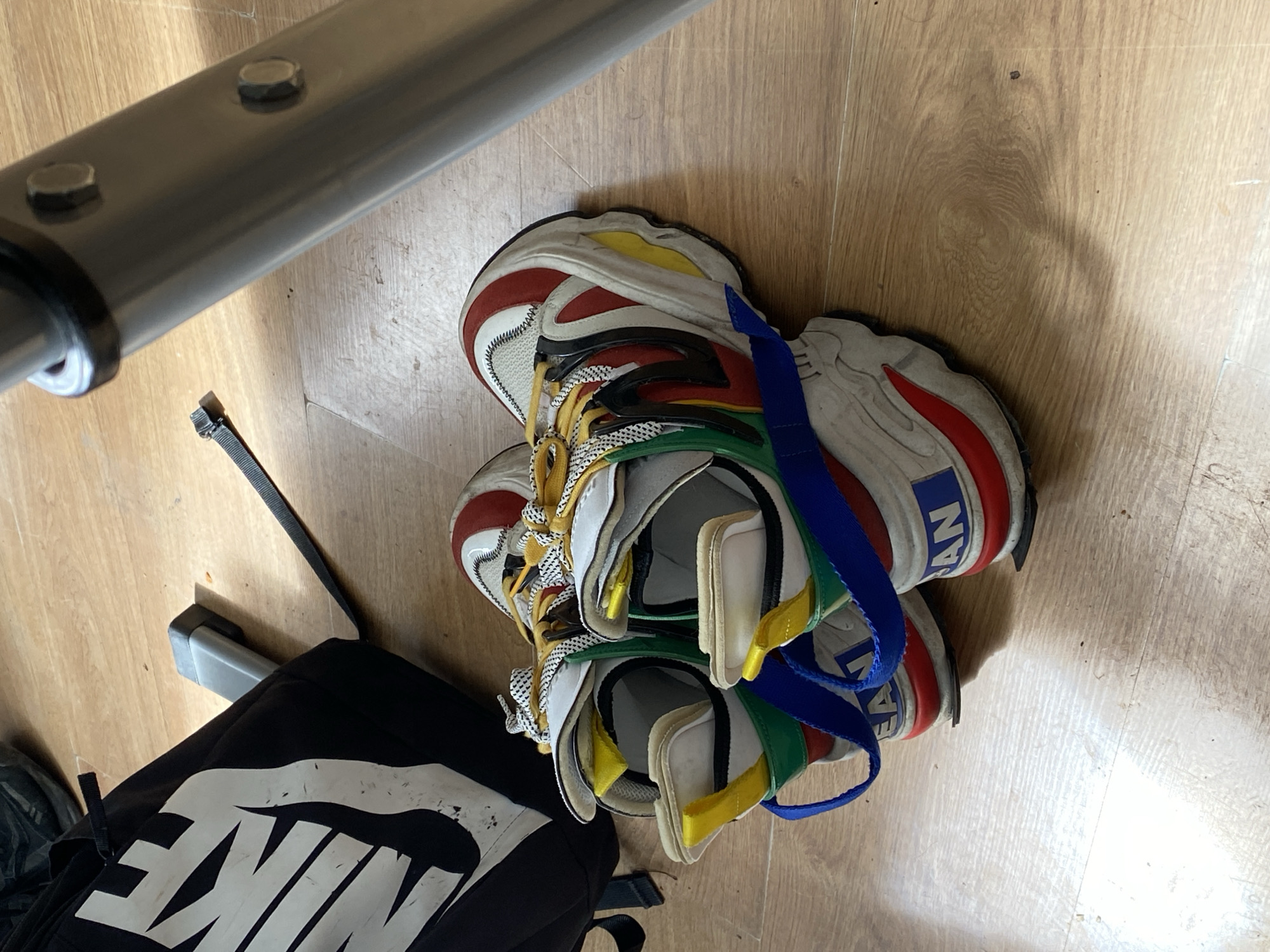 The image shows a pair of colorful sneakers stacked on top of each other on a wooden floor. A black backpack with a large white Nike logo is partially visible in the lower-left corner. A metal bar with bolts is in the upper-left corner, and a thin black strap lies near the backpack. The context of Varna, Bulgaria, is not directly evident within the visual content of the image.