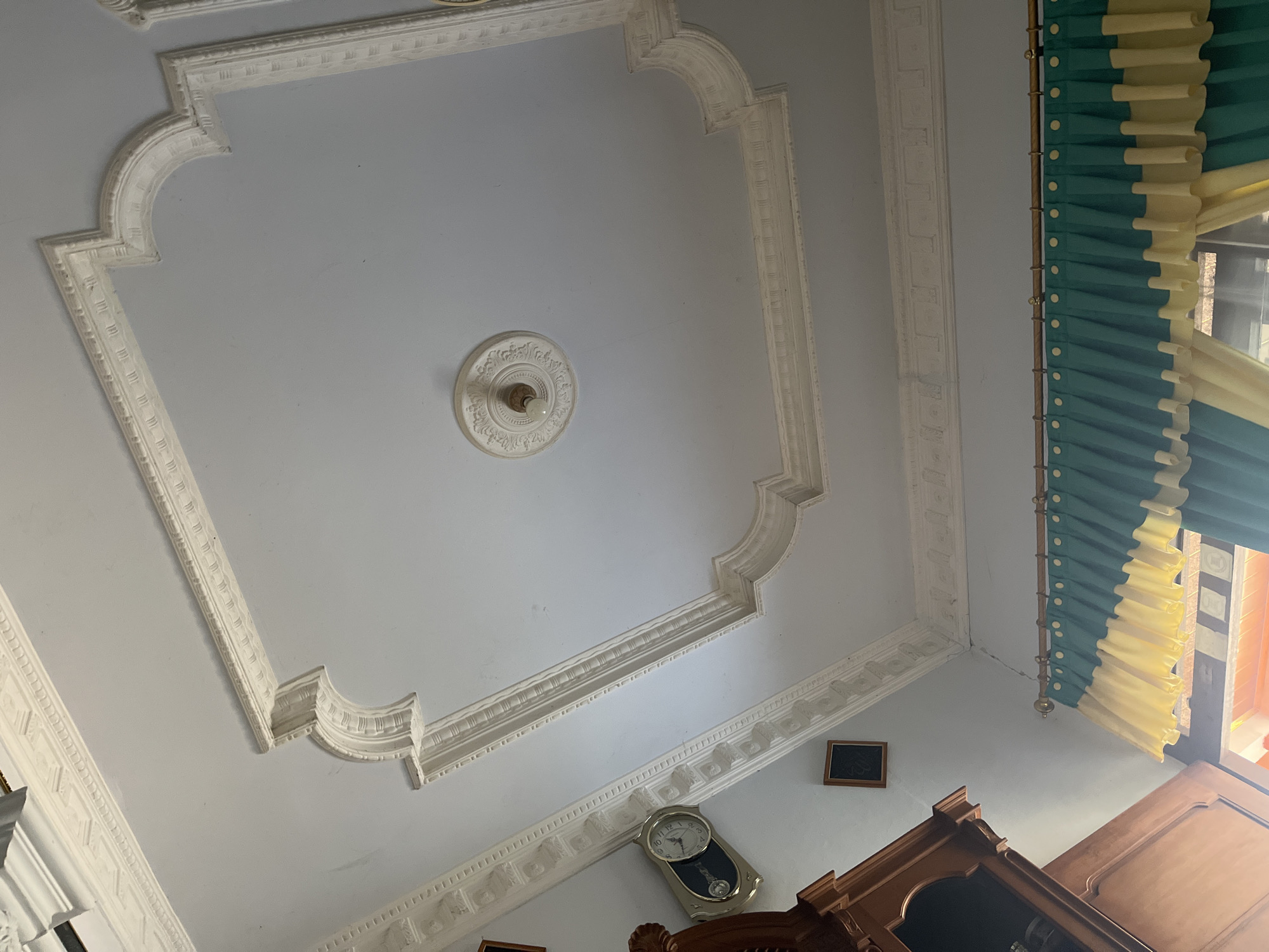 The image shows the interior of a room with a white, decorated ceiling. Intricate molding frames a large rectangular section of the ceiling, with curved corners and detailed patterns along the edges. In the center of the room, a decorative ceiling rose surrounds a light fixture with a visible light bulb.
On the left side of the image, a portion of a wooden cabinet or piece of furniture is visible. Mounted on the wall next to it is a clock with a silver frame and a black face, showing the time to be approximately 10:10. A small, framed picture or plaque is also visible on the wall above the clock.
The lower part of the image shows a window with curtains. The curtains are a combination of teal and yellow fabric, hung on a gold-colored rod. The teal curtains are pleated and gathered, with yellow fabric layered beneath them, creating a decorative effect. The sunlight filtering through the window suggests it is daytime.
The environment appears to be a residential interior, possibly a living room or dining area, located in Glagah Kulon, Indonesia. The overall impression is one of a well-decorated and maintained indoor space.