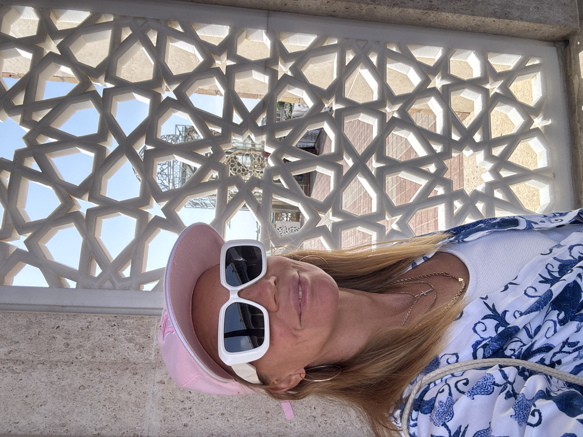 The image shows a woman in a pink baseball cap and white sunglasses, standing in front of a decorative white stone wall with geometric star patterns. The wall appears to be part of a building or structure in Samarkand City, Uzbekistan, as architectural details visible through the wall's openings suggest Central Asian design. The woman is wearing a white top with a blue floral patterned overlay. She also has a gold necklace and gold hoop earrings. The photograph is taken at an angle.