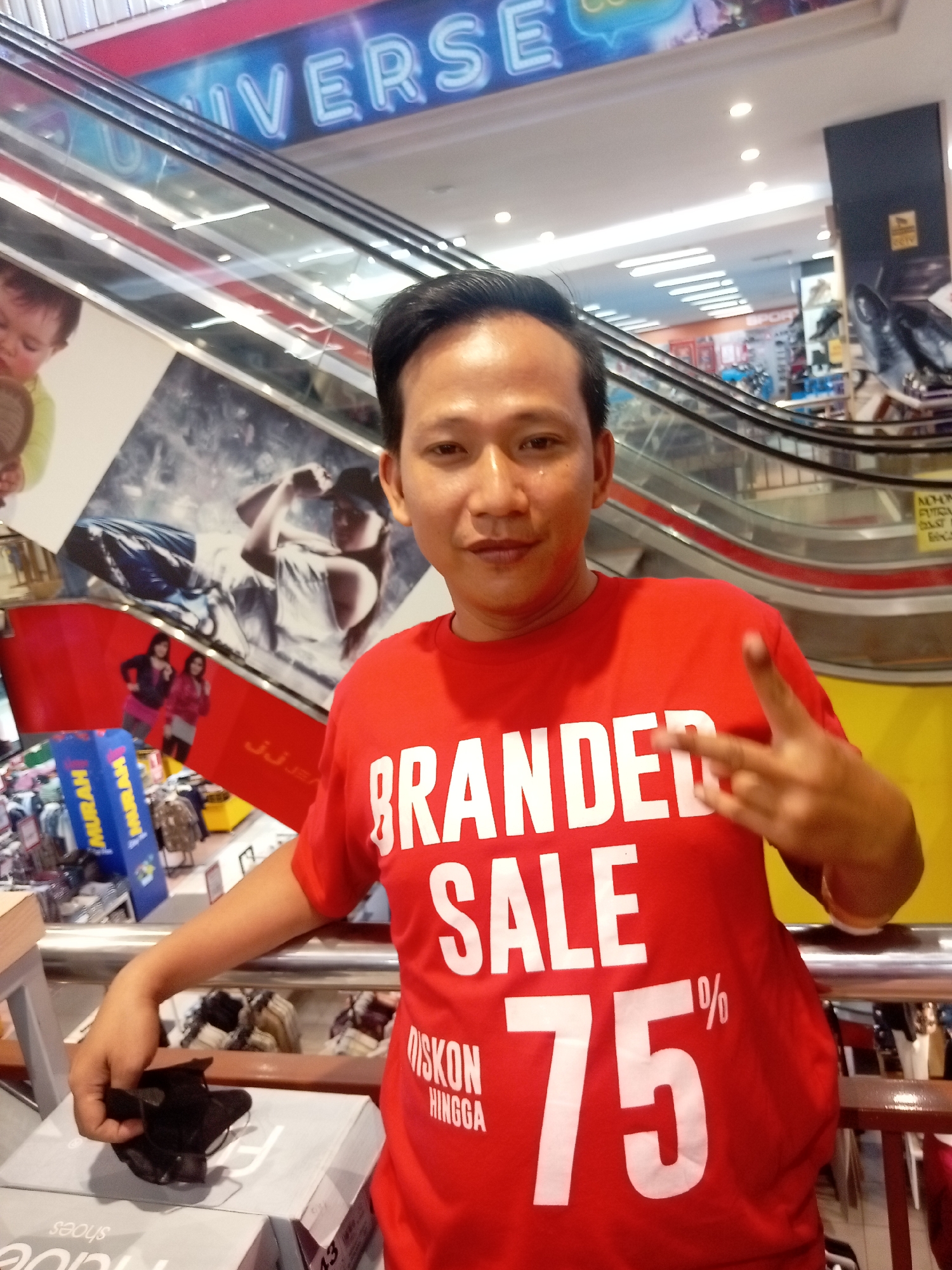 A man in a red t-shirt with "BRANDED SALE 75%" printed on it is posing for a picture in a shopping mall. He is making a peace sign with his right hand. The background shows escalators, shop signs, and advertisements. One sign above the escalator reads "UNIVERS". The man is holding a black object in his left hand, possibly a wallet or a small bag. The scene appears to be in Batam, Indonesia, likely in a shopping mall like Batam City Square (BCS) or Mega Mall, given the presence of the "UNIVERS" signage which could refer to Universal Studios or a similar branding often found in large retail spaces in Indonesia.