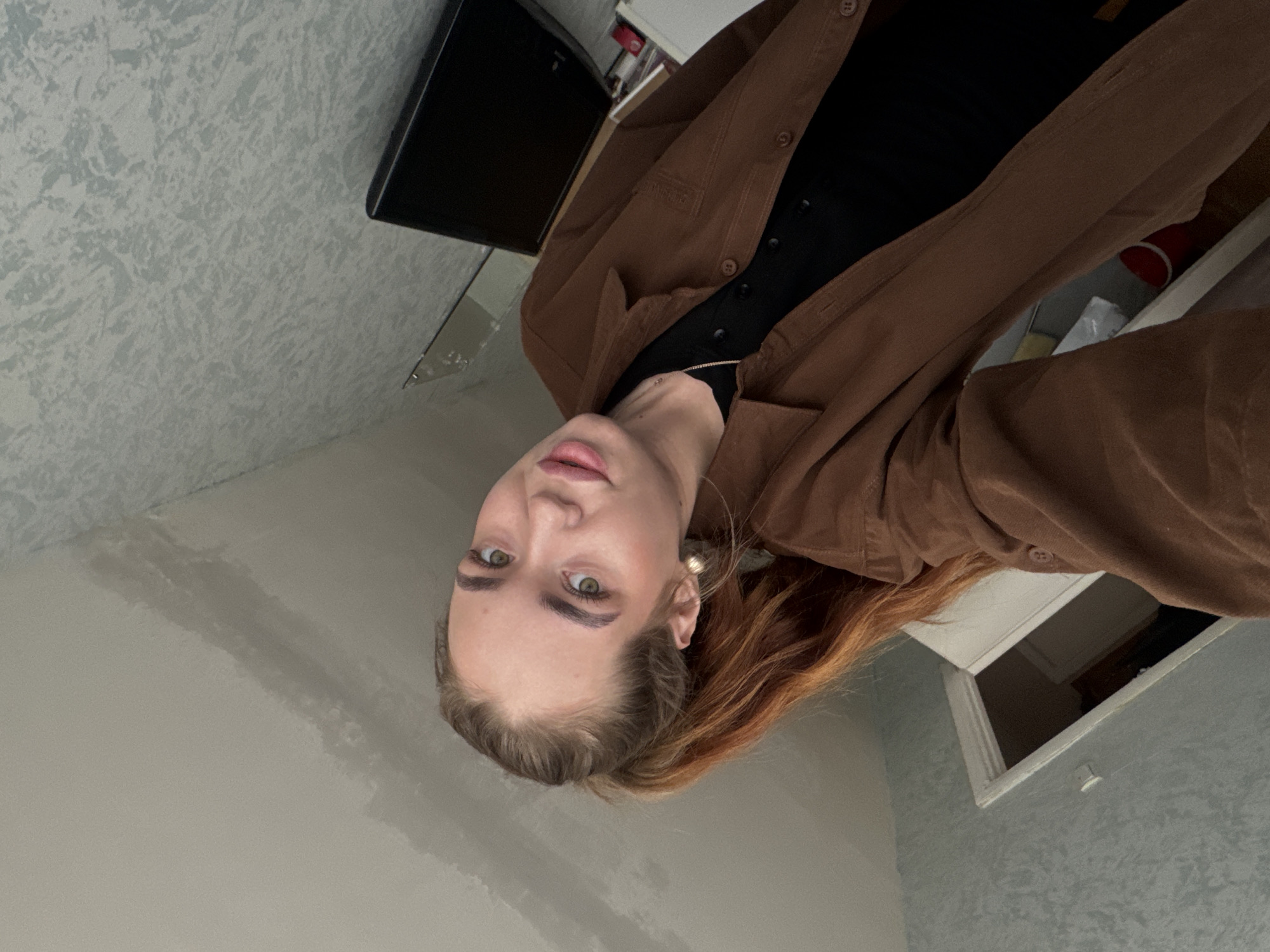 A young woman with light skin and green eyes is centered in the image. She wears a black top and a brown button-down jacket. Her hair is a mix of brown and reddish tones. Behind her, a wall is visible with a light blue patterned texture. A dark object resembling a small television or monitor is partially seen in the upper left corner. An open window is visible in the upper right. The image location is Chaikovsky, Russia.