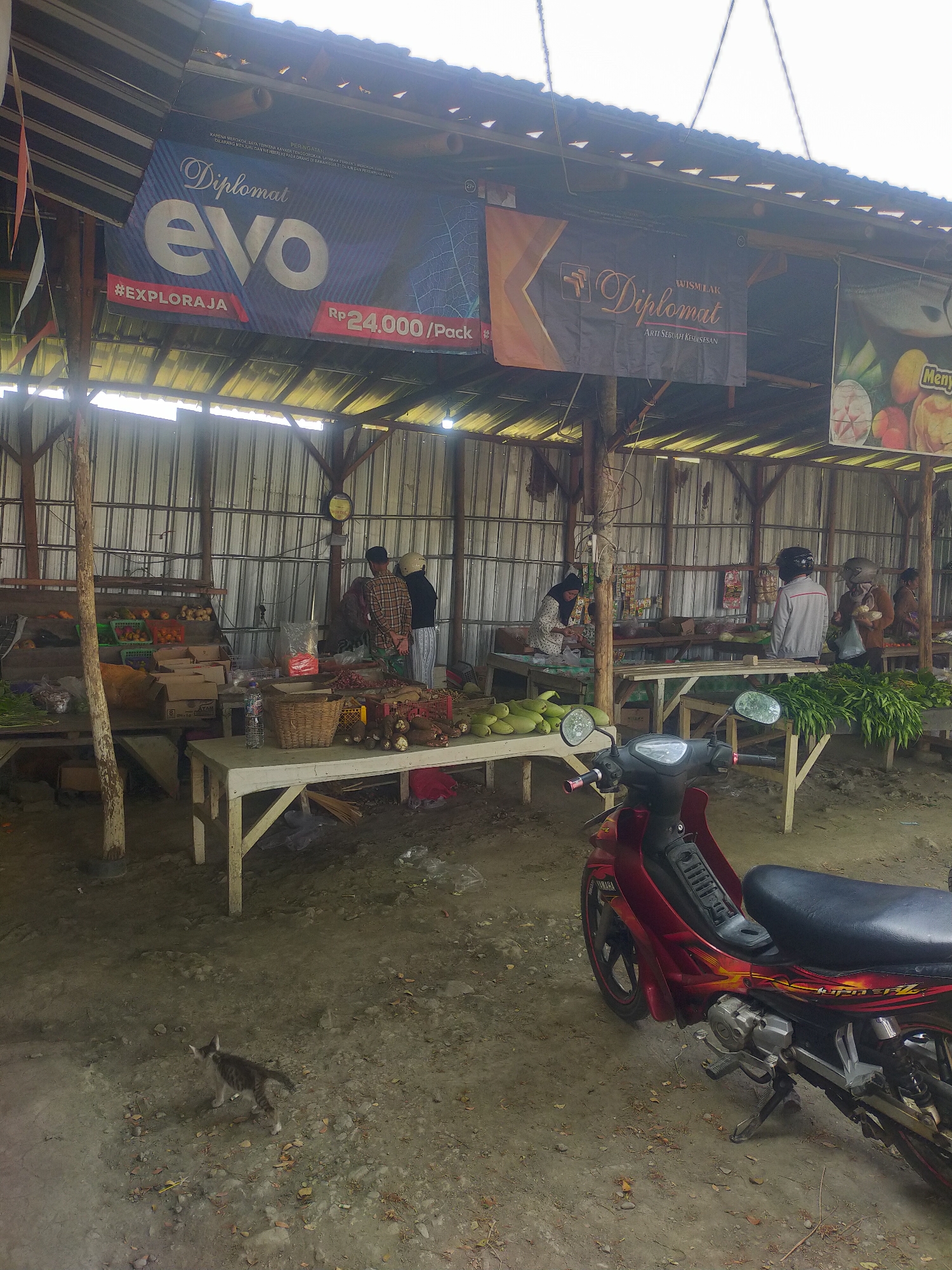 The image depicts an outdoor market scene in Kalisapu, Indonesia, under a covered structure. In the foreground, a red motorcycle is parked on a dirt ground. The market stall area features wooden tables laden with produce such as leafy greens, gourds, and various vegetables. Several people are visible in the market, some attending to the stalls and others appearing to be customers. The stalls are organized with baskets and boxes holding the merchandise. Hanging banners display advertisements, one of which features "Diplomat evo" and a price. The overall environment suggests a typical rural or semi-urban market setting. A small grey and white kitten is also present in the foreground, walking on the dirt.