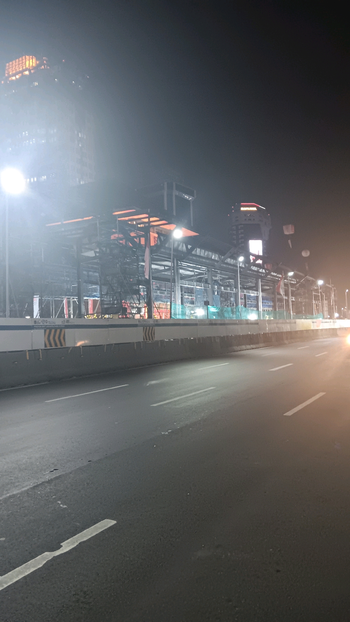 An nighttime, low-angle, outdoor vertical shot shows a road in front of a construction site in Central Jakarta, Indonesia. The road is dark asphalt with white lane markers. A concrete barrier with yellow and black chevron markings separates the road from the construction area. The construction site is illuminated with bright lights, revealing scaffolding, steel beams, and what appears to be building materials. In the background, a tall, modern building with glowing windows rises into the foggy night sky. The overall atmosphere is hazy, possibly due to pollution or fog.