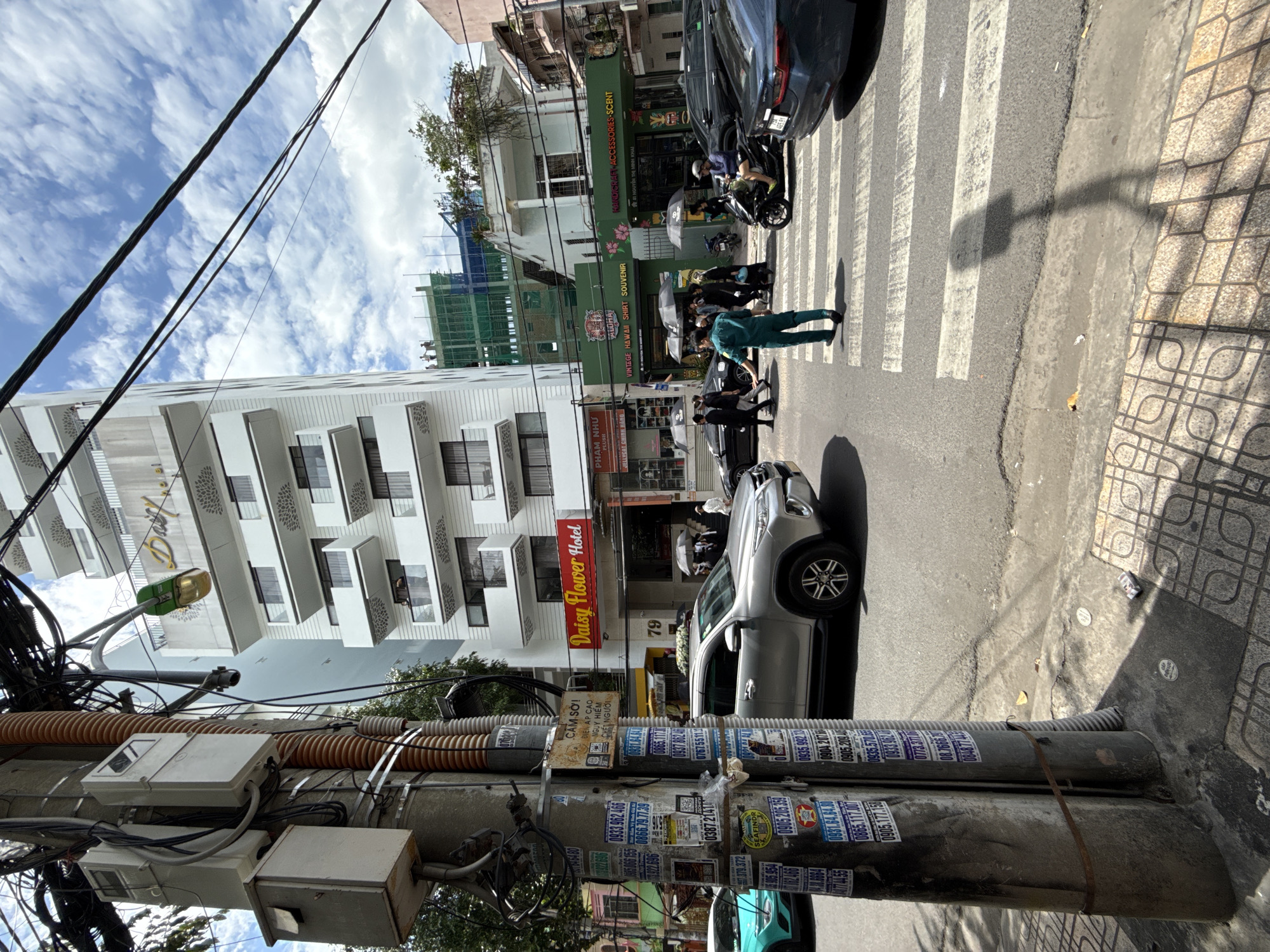 In Nha Trang, Vietnam, a street scene unfolds at a crosswalk. A silver SUV is stopped on the right side of the street, partially obscuring the view of a group of pedestrians crossing. One pedestrian, dressed in a teal uniform, is in the process of crossing. Behind the SUV, a dark-colored sedan is visible, also stopped. On the left side of the image, a teal car is parked. Overhead, a tangle of electrical wires and a street light are prominent. In the background, a tall, white, multi-story building with balconies displays a sign for "Daisy Flower Hotel". Adjacent to the hotel, a green storefront with the word "Aloha" and signage for "VINTAGE HAWAIIAN SHIRT SOUVENIR" and "HANDICRAFT ACCESSORIES - SCENT" is visible. The sky above is cloudy with patches of blue. The street is paved, with a marked crosswalk. A utility pole on the left is covered in numerous advertisements.
