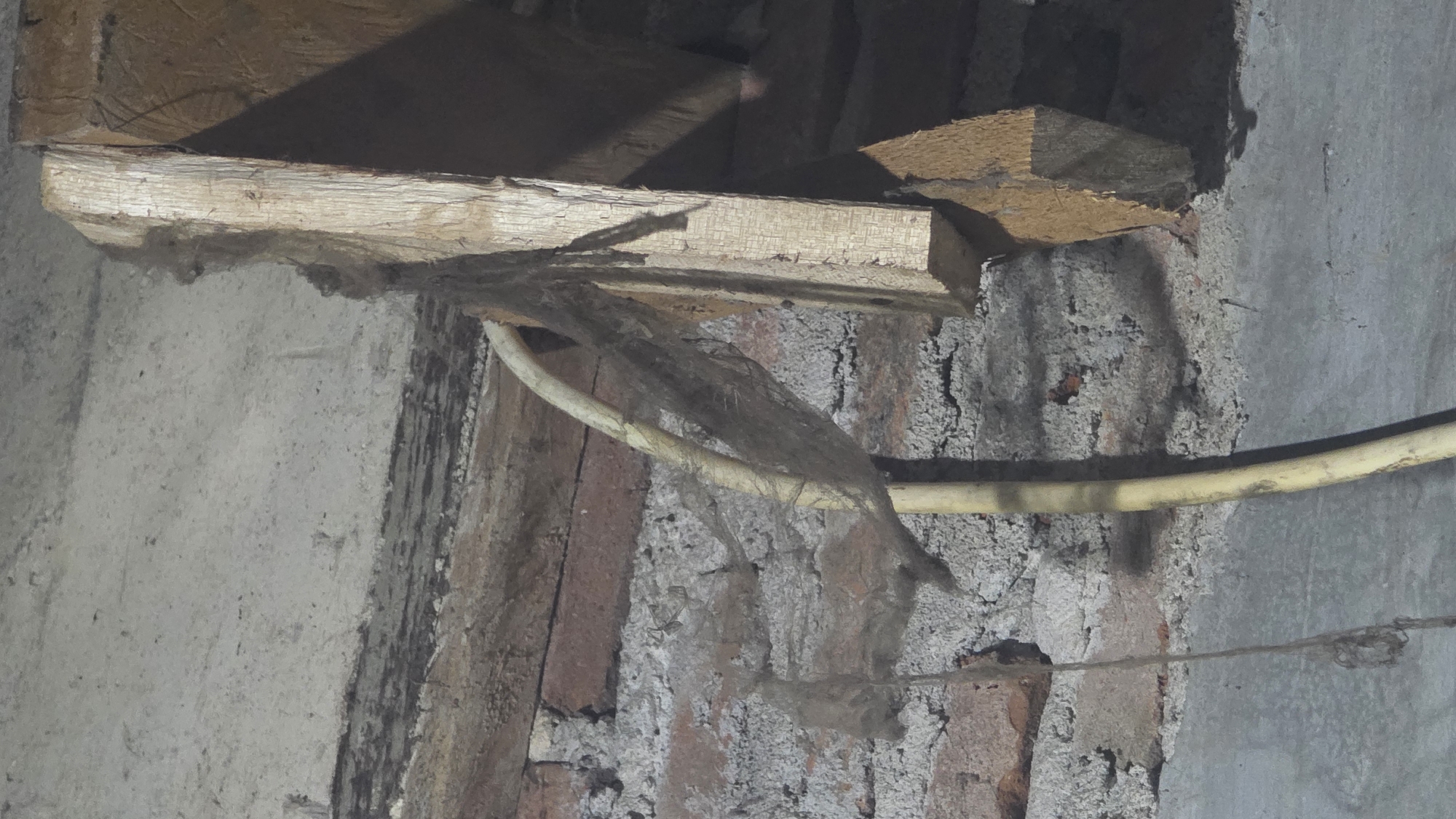 The image depicts a section of exposed interior wall in Lhokseumawe, Indonesia. The wall construction is a combination of brick and concrete. Sections of the brick are exposed, with crumbling mortar in between. A horizontal yellow electrical conduit runs across the face of the wall, partially obscured by cobwebs. Above, unfinished wooden beams are visible, partially embedded within the wall structure. Dust and debris cling to the wood.