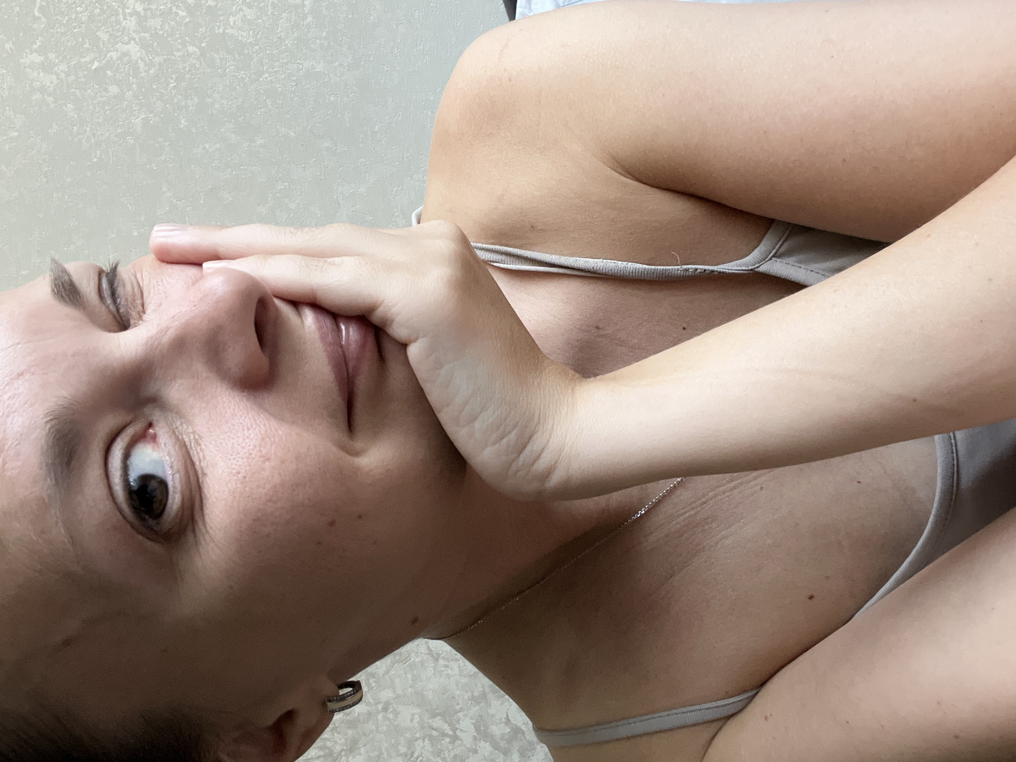 A close-up, profile view of a woman winking and resting her chin on her hand. She is wearing a gray camisole and a silver necklace. She has on a small silver hoop earring in her left ear. The background is a textured, neutral-colored wall. No specific location in Tolyatti, Russia, can be identified.