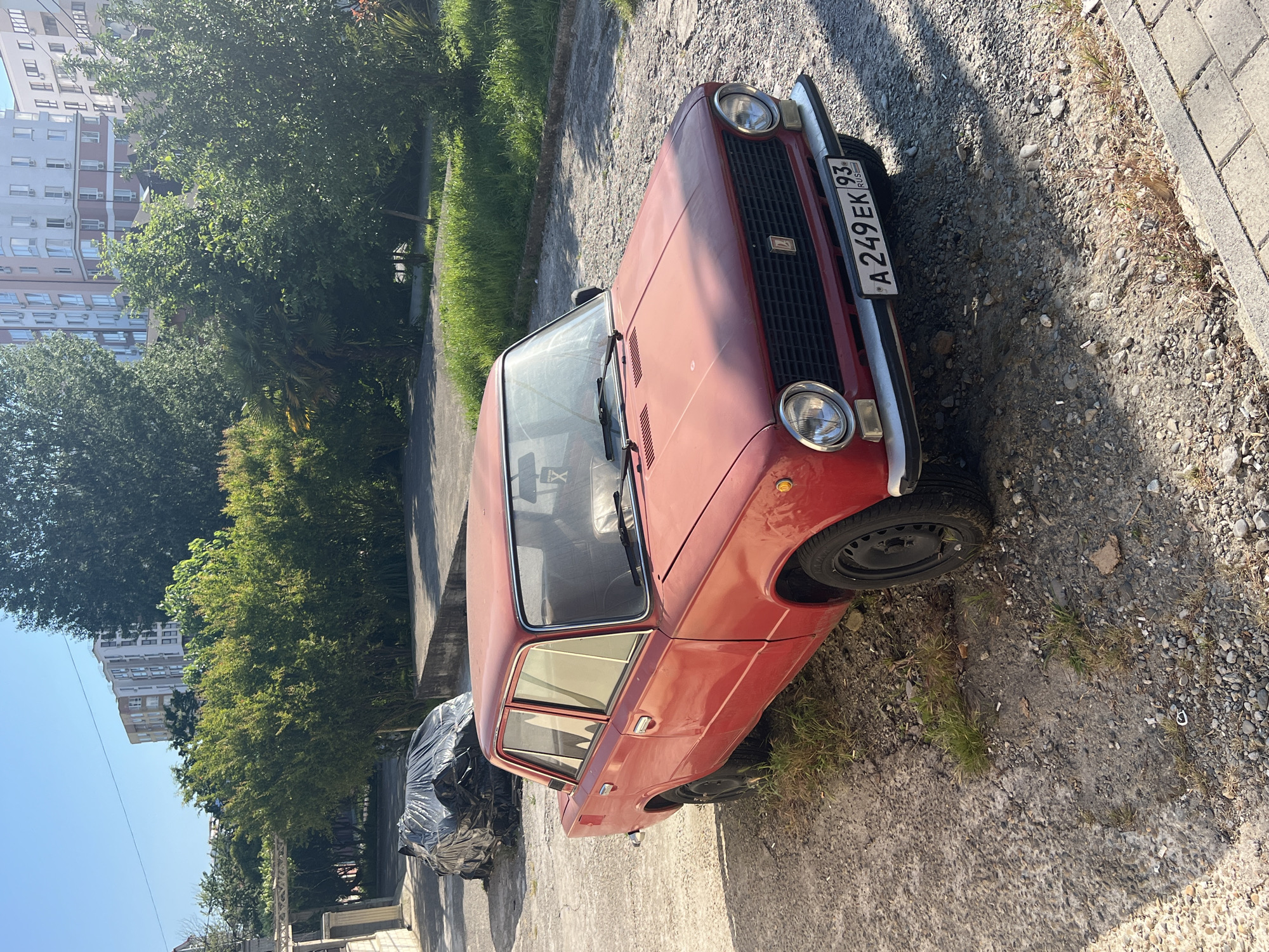 A red, older model sedan is parked on a gravel and asphalt surface in a sunny outdoor setting. The car, which appears to be a Lada, has a license plate that reads "A 249 EK 93". In the background, there are lush green trees and bushes, along with a modern multi-story building. To the left of the car, another vehicle is covered with a dark tarp. The ground is a mix of small rocks, dirt, and sparse dry grass. A paved sidewalk is visible in the lower right corner of the image. The sky is clear and blue, indicating a bright day. This scene is consistent with a residential or less maintained area within the city of Sochi, Russia.