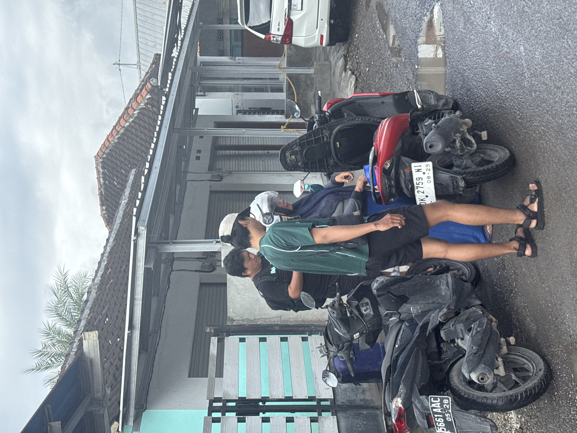 The scene shows three people in Kampung Toyapakeh, Indonesia, leaning on two parked motorbikes. One person is wearing a white helmet, a dark blue jacket, and sandals. Another person is wearing a green shirt, black shorts, and sandals. The third person is wearing a black shirt. The motorbikes are parked on a paved street. One is black, and the other is red. A white car is parked nearby. The street is lined with buildings. The sky is overcast.