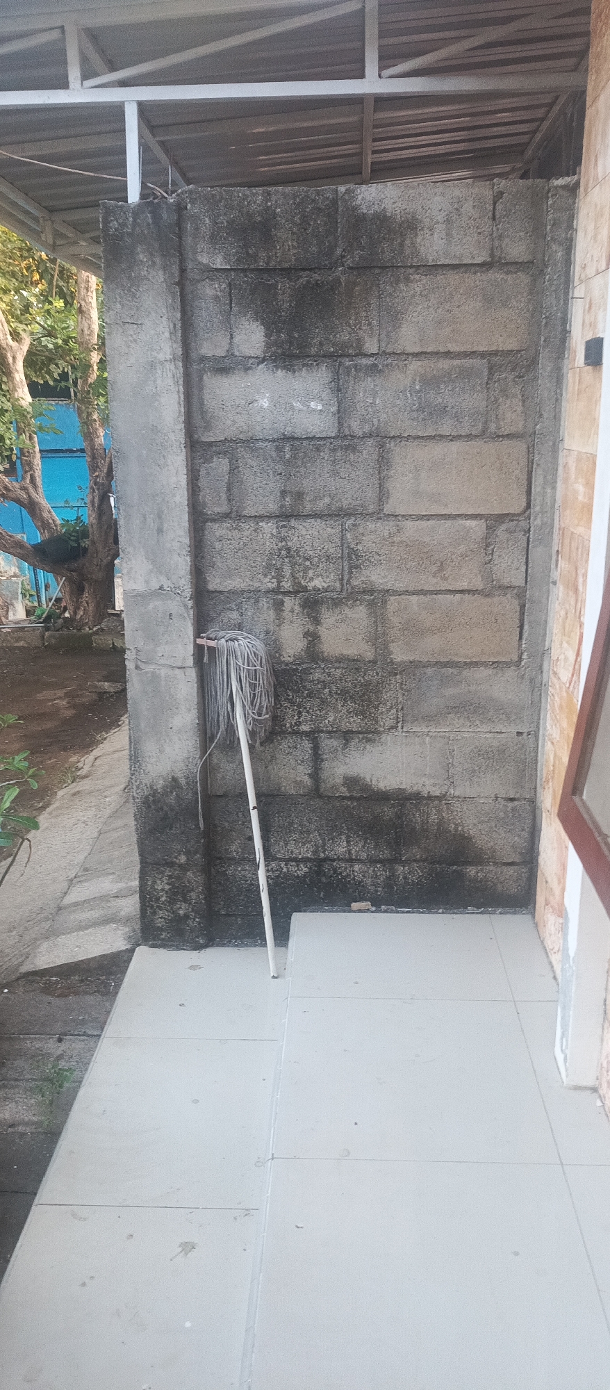 The image depicts an outdoor scene in Jatisari, Indonesia, featuring a rough, grey cinder block wall as the central element. A white mop with grey strings is leaning against the wall, positioned to the left of the wall's center. The wall shows signs of weathering and staining, with darker patches suggesting moisture or grime. To the left of the wall, a concrete pillar stands, and beyond that, a dirt path is visible, leading to a tree with exposed branches. A portion of a metal roof structure is visible above the wall. The foreground consists of light-colored tiled flooring. The overall impression is one of a utilitarian outdoor space, possibly a service area or alleyway, with evidence of recent cleaning or a mop left in place.