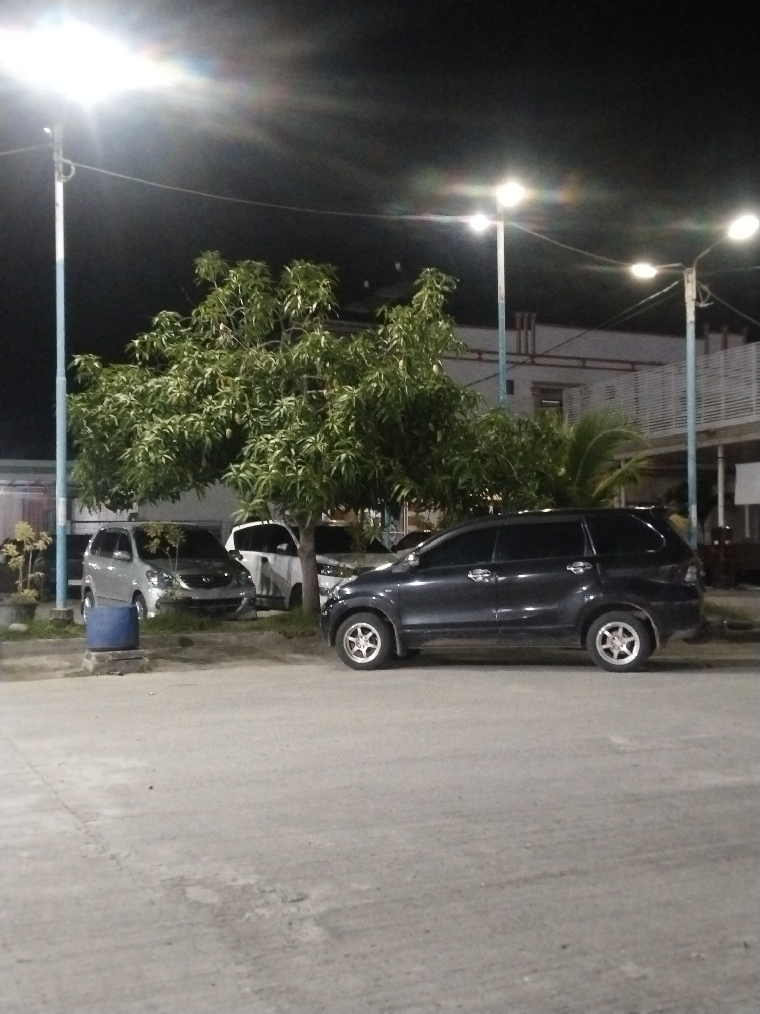 At night, a dark-colored SUV is parked on a paved surface in front of a large, leafy tree. Behind the tree, several other vehicles are visible, including a silver minivan and a white car. A building with an elevated walkway and railings is situated in the background. Streetlights illuminate the scene, casting a glow on the surroundings. The location appears to be a parking area in Keuniree, Indonesia.