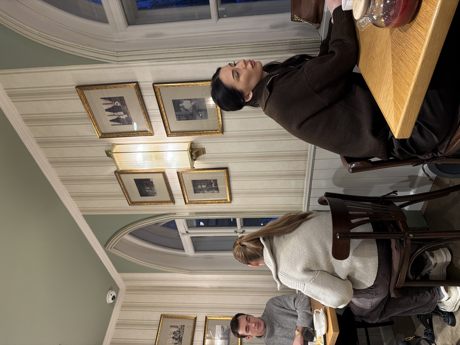 Three people are seated at a light-colored wooden table. One person wears a brown sweater and sits to the left, another person wears a light gray sweater, and the third person wears a gray sweater and sits in the bottom middle of the frame. The dining area has striped wallpaper, windows with arched tops, and framed pictures on the walls. A rectangular lighting fixture is mounted on the wall. A security camera is visible.