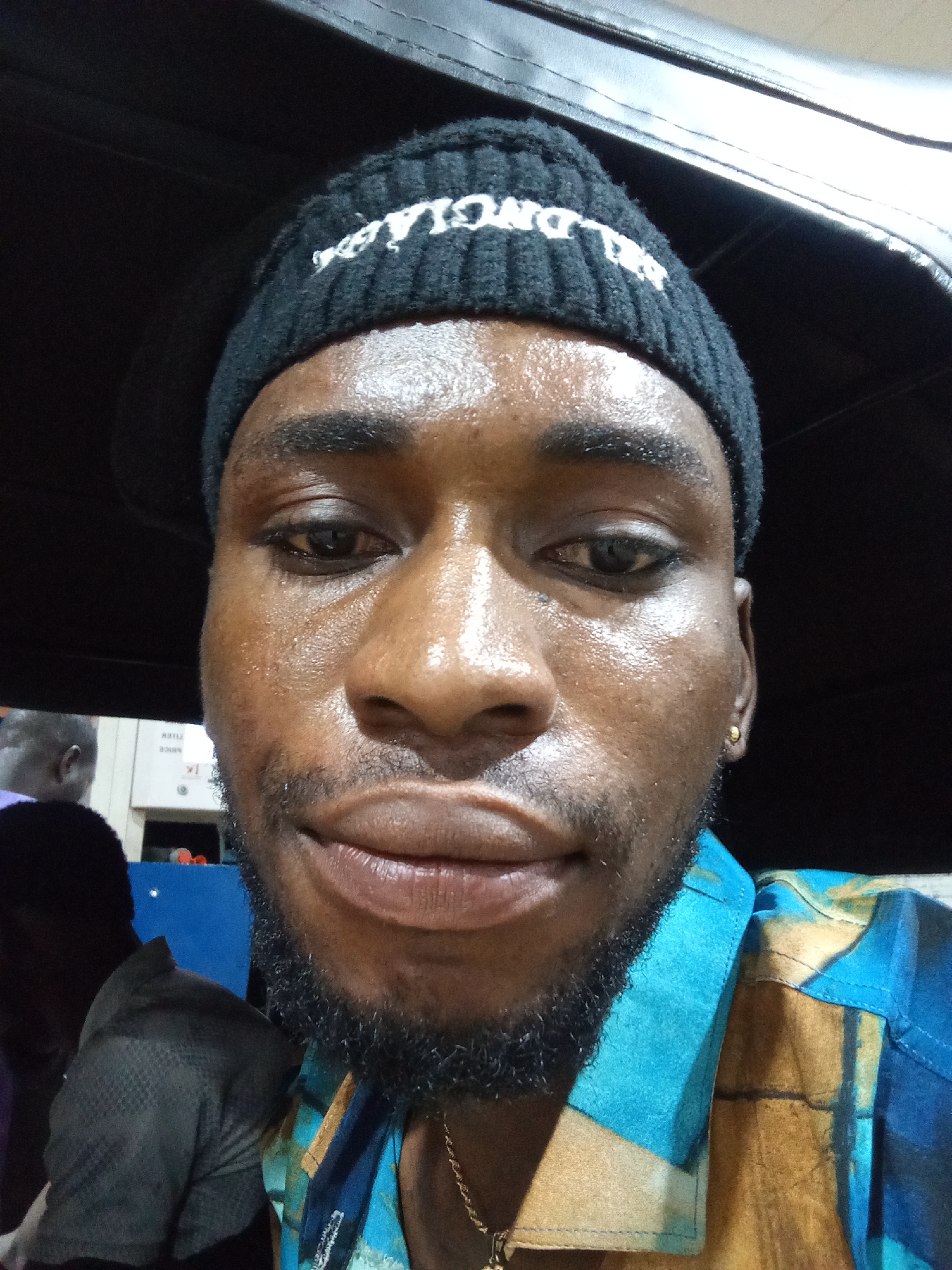A close-up selfie of a Black man wearing a black beanie with white lettering and a blue and gold patterned shirt is shown. He is looking directly at the camera. The background is dark, with some blurred figures visible on the left, suggesting an indoor setting, possibly a public transport vehicle given the roof-like structure visible above. The man's face is slightly sweaty, and he has a goatee and a gold earring. The setting is implied to be within the city of Lokoja, Nigeria, though no specific landmarks are visible in the image to confirm this.