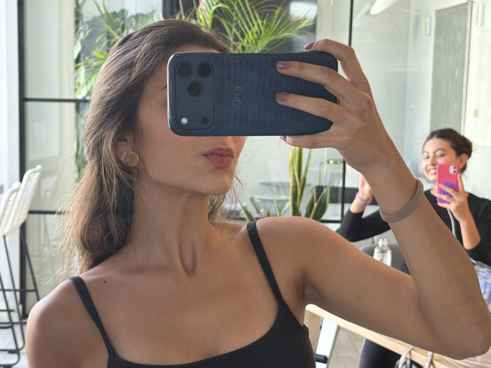 A mirror selfie captures two females in an indoor setting, identified as being in Miami, United States of America. The primary subject, in the foreground, is a female with long brown hair, wearing a black spaghetti strap top and a gold circular earring with an etched design. She holds a smartphone with a dark blue, textured case featuring a triple camera array, obscuring her upper face; her lower face shows pursed lips with light pink lipstick. A light brown elastic band is visible on her right wrist. Reflected in the mirror in the background, a second female with dark hair pulled back is smiling brightly, holding a bright pink smartphone with a purple case as if taking her own selfie. She wears a black long-sleeved top and a pink elastic wristband. The reflected background includes a tall green plant, a black-framed glass partition, a partially visible white wicker-style bar stool, and a clear water bottle, suggesting a modern, well-lit interior space. Both individuals are engaged in the act of taking self-portraits using their smartphones and the reflective surface.