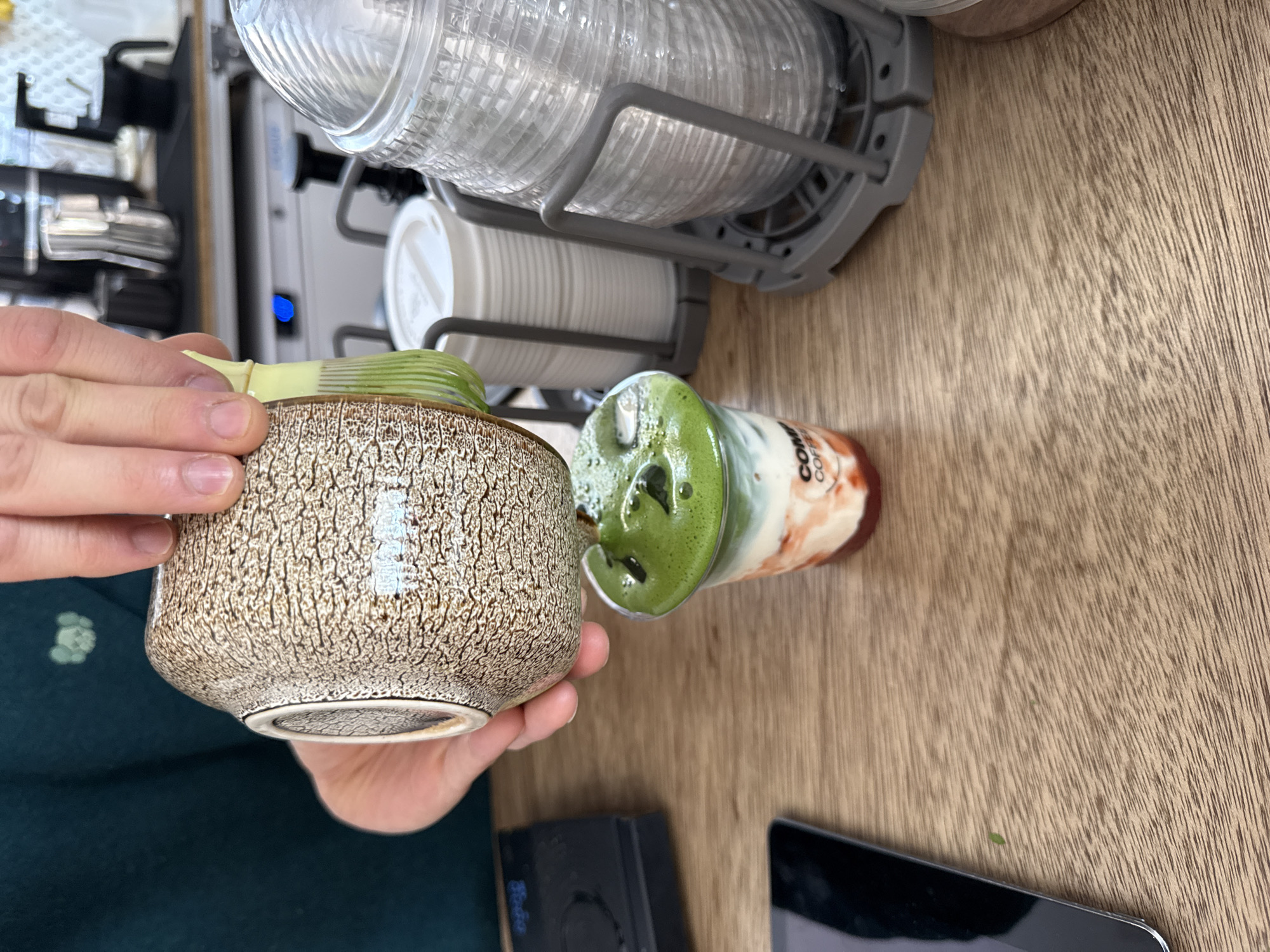A top-down, slightly angled shot captures a hands-on moment at a wooden counter, likely in a cafe or kitchen in London, United Kingdom. The central focus is a person's hands holding a distinctive ceramic bowl with a textured, mottled brown and beige pattern. Inside the bowl, the upper part of a light-colored bamboo whisk (chasen) is visible, suggesting the preparation of matcha.
To the right of the bowl, a clear plastic cup sits on the counter, containing a layered beverage. The drink features a prominent green upper layer, possibly matcha, with a frothy top and small darker specks, resting above a reddish-brown liquid at the bottom. Partial text "COFF" is visible on the side of the cup.
In the background, a metal rack organizes stacks of clear plastic cups and white paper cups, indicating a beverage service area. A machine with a blue light is faintly visible behind the cups. The countertop itself is made of light-colored wood with a visible grain. In the lower right corner, a black electronic device and a portion of a tablet or smartphone screen are visible. The person holding the bowl is wearing dark green attire. The bright lighting suggests it is daytime.