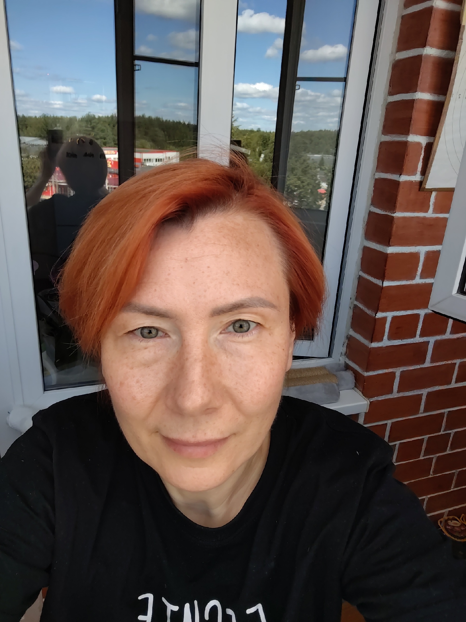 A red-haired woman with freckles takes a selfie on a balcony in Konakovo, Russia. The woman is wearing a black sweatshirt. Behind her, through a window, is a reflection of the woman holding her phone up to take the picture. The background outside the window shows a clear blue sky with clouds, trees, and buildings. To the right of the woman is a brick wall.