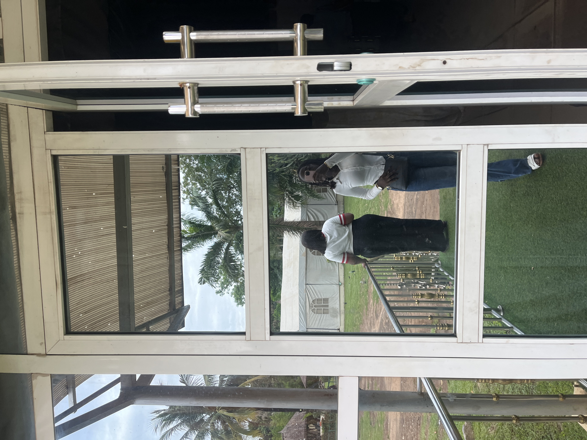 The scene is set in Galadimawa Village, Nigeria, observed from an interior through a white-framed glass door and an adjacent fixed window panel. The glass surfaces exhibit reflections of the interior space and occupants, concurrently allowing a view of the exterior environment.
On the right side of the image, the white-framed glass door is partially ajar, revealing a dark interior. It features two prominent, vertically oriented silver-colored bar handles. The glass panels are streaked with minor smudges and dirt.
Reflected in the right-most glass panel, a person with dark skin and dark hair, styled in dreadlocks or braids, is visible. This individual is taking a selfie, holding a light-colored smartphone to their face, obscuring their upper facial features. They are wearing a white, long-sleeved, collared shirt, blue jeans, and light-colored sandals. A dark shoulder bag is slung across their body, and a watch is visible on their left wrist.
Reflected in the left glass panel and seen directly through it, another individual is positioned with their back to the viewer, leaning against a metallic railing. This person has dark hair and is wearing a white t-shirt with red striped accents on the sleeves and a long black skirt or dress.
The interior foreground, visible through the lower glass panels and in reflection, features green artificial turf. The railing, made of silver-colored metal with ornate gold-colored balusters, is visible both directly and as a reflection.
The exterior view beyond the glass displays an overcast or partly cloudy sky. The landscape is characterized by abundant green foliage, including several tall palm trees. In the mid-ground, a large white event tent with arched window cutouts is clearly visible on a grassy, somewhat dirt-patched area. Several small dark birds are scattered on the ground near the tent. In the far left background, partially obscured by trees, are smaller, rustic structures with thatched roofs.