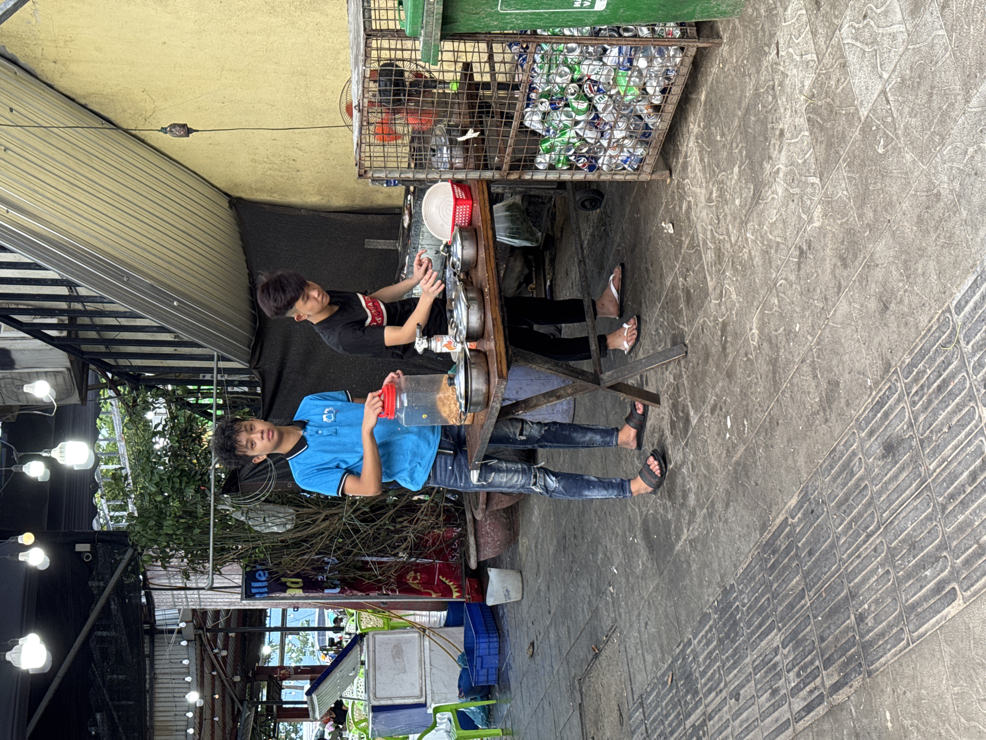 Two young males are standing at a makeshift food stall on a paved outdoor area. The stall is set up on a wooden table, with various containers of food and utensils. The male on the left, wearing a blue polo shirt and ripped jeans, stands behind a large plastic container filled with food. The male on the right, wearing a black t-shirt, is actively preparing food at the table. Behind them, a metal cage is filled with empty aluminum cans, and a green trash bin is visible to the right. The background shows a mix of covered structures, vegetation, and other stalls, suggesting a busy street market or food court. The scene appears to be in an urban setting, likely a street or market in Đà Nẵng, Vietnam, given the context.