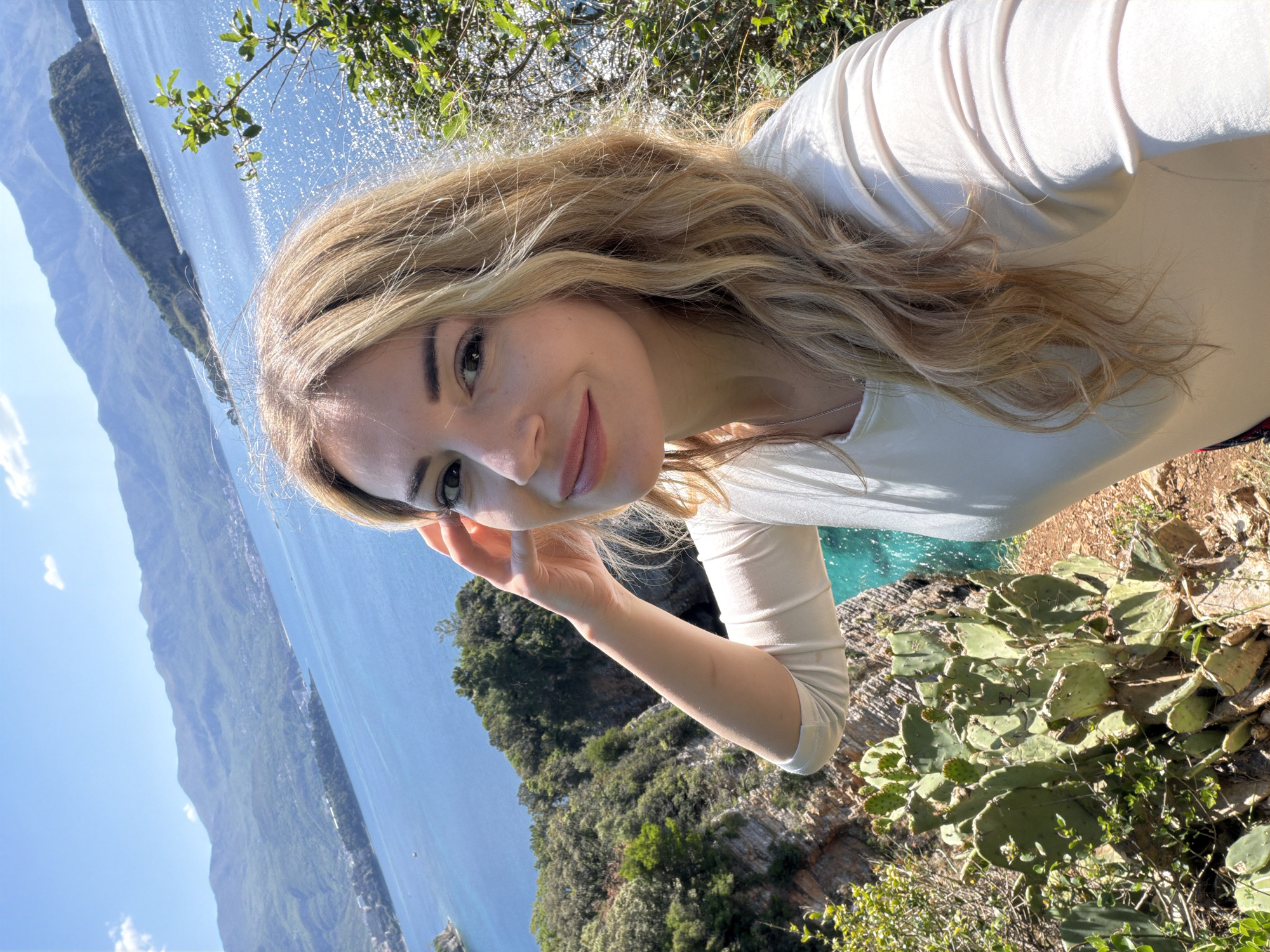 A selfie of a blonde woman with long, wavy hair, wearing a white, long-sleeved shirt. She is smiling slightly, with her right hand raised near her face. In the background is the Adriatic Sea, a mountainous coastline, green vegetation, and some cacti. The location is likely a coastal area near Budva, Montenegro, given the landscape and seascape. The turquoise-colored water is notable.