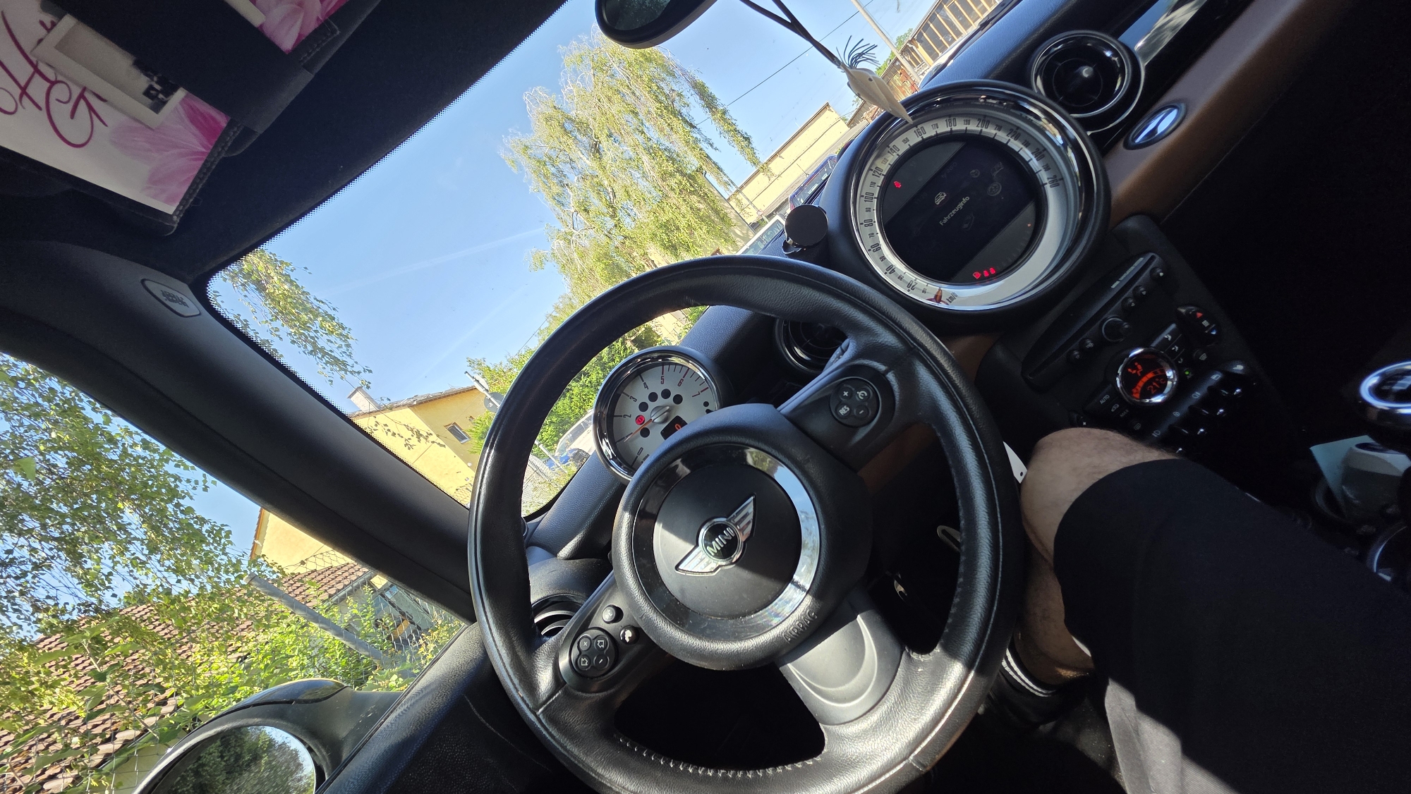 The image shows the interior of a Mini Cooper car, looking out through the windshield. The steering wheel is visible in the foreground, with the Mini logo at its center. A person's leg and knee, clad in black shorts, are visible on the left side of the image. The car's dashboard displays various gauges and controls. Outside the car, trees and a yellow building are visible under a clear blue sky. The scene suggests a sunny day in an outdoor environment, possibly a residential area.