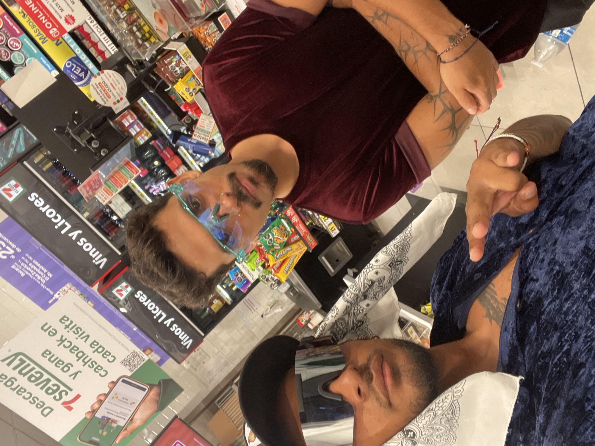 The image shows two men indoors, likely within a convenience store, possibly in Playa del Carmen, Mexico, given the Spanish text on the signs.
The first man is wearing a maroon, velvet-like shirt and has visible arm tattoos. The second man is wearing a blue, velvet-like shirt and a patterned white bandana. Both men appear to be in a seated or reclined position, possibly taking a selfie or group photo.
The background includes shelves filled with items for sale, such as snacks, beverages, and tobacco products. There are signs indicating discounts and promotions, as well as signage for "Vinos y Licores" (wines and liquors). Other discernible items include a cash register or payment terminal, and promotional materials related to a mobile app called "7sevenly" that offers cashback.