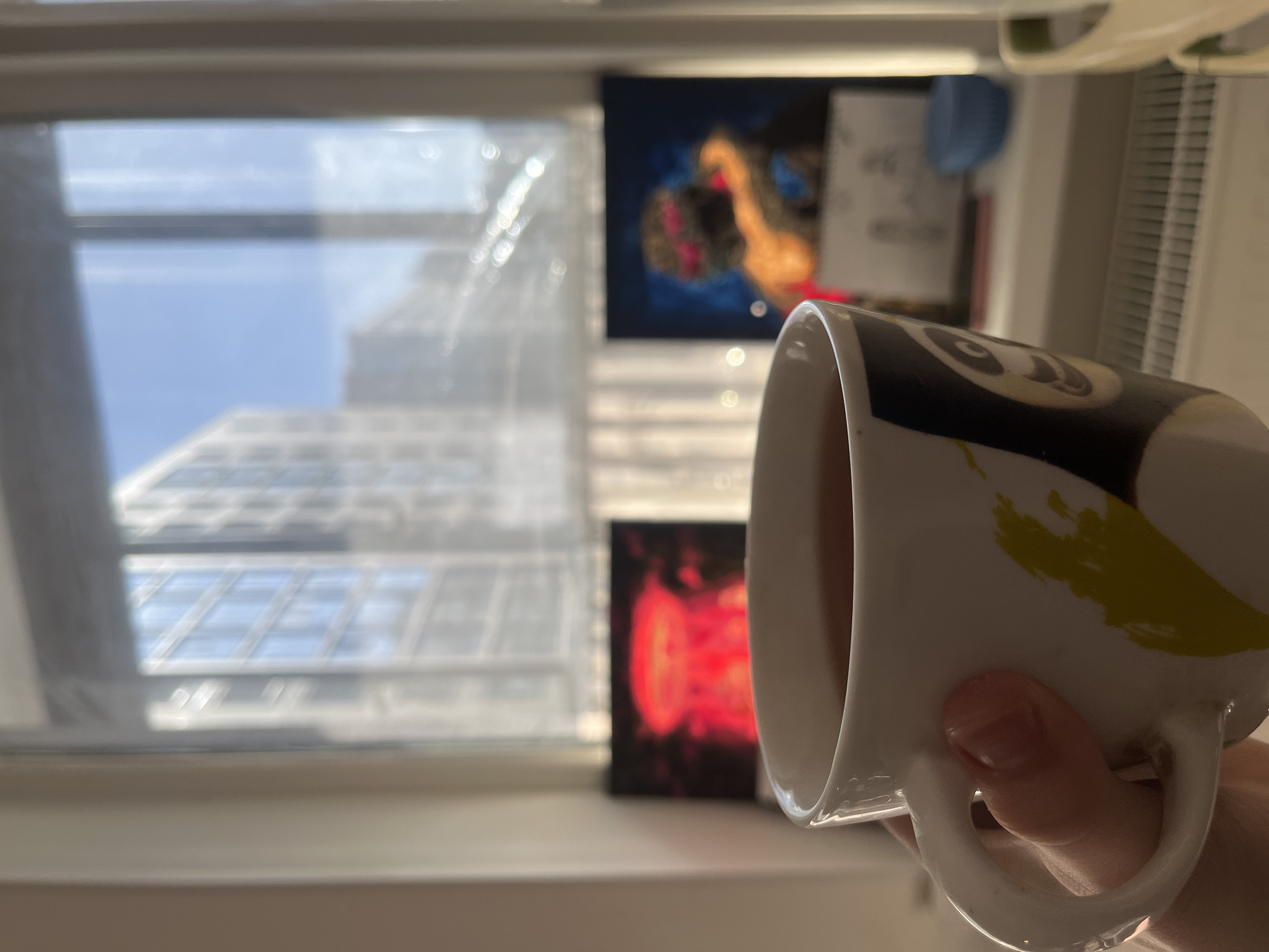 A person holds a white ceramic mug with a black and yellow design in front of a window. The mug is filled with a light-colored liquid. Outside the window, a blurry view of a tall building with many windows is visible against a blue sky. Two paintings hang on the wall behind the window. The setting appears to be indoors, possibly in an apartment or office. It is impossible to ascertain the location as Saint Petersburg, Russia from the image alone.