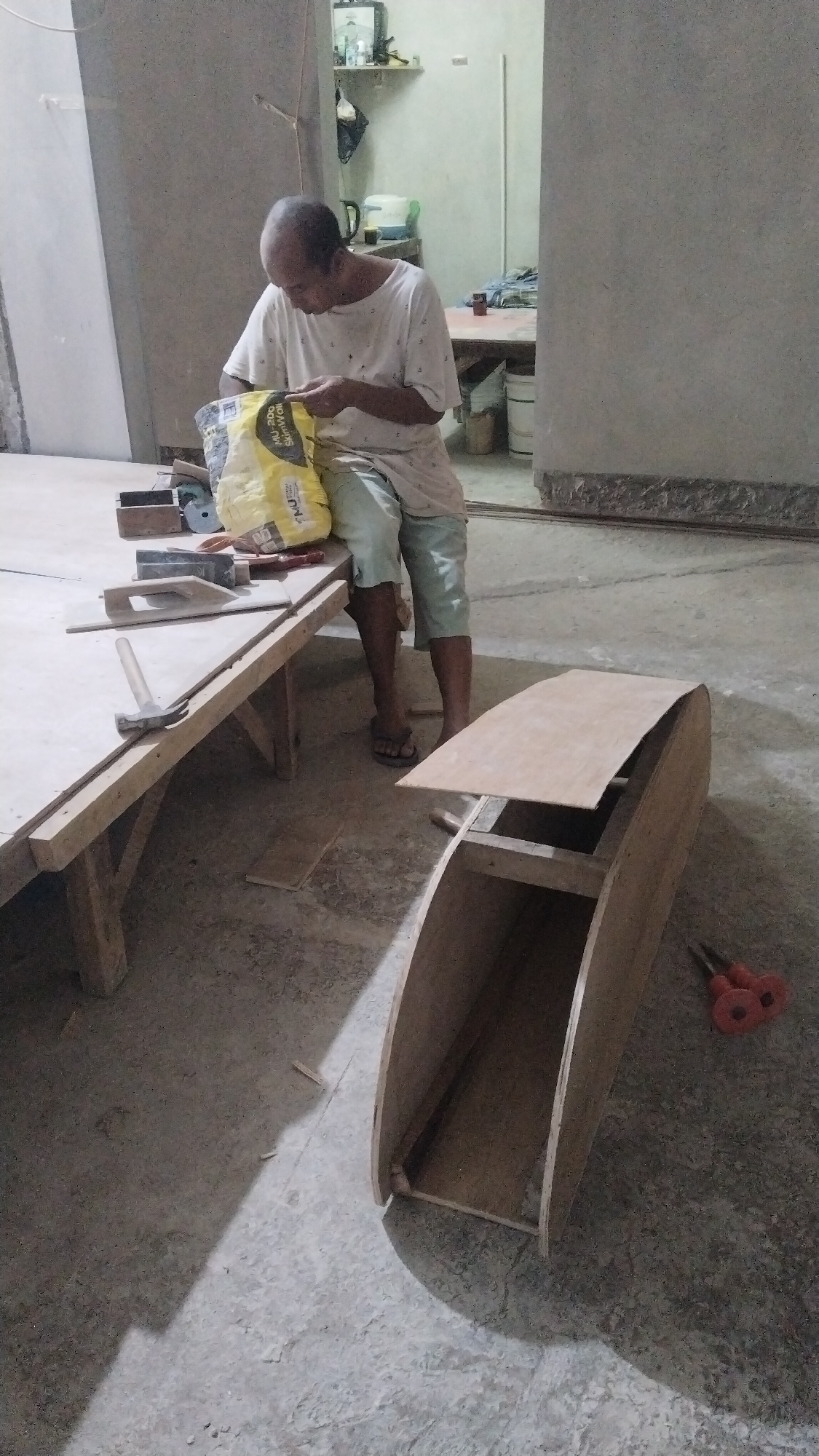 An overhead shot shows a bald, middle-aged man wearing a white t-shirt and light blue shorts. He is kneeling on the floor in what appears to be a workshop or construction site. He is holding a yellow bag with text on it and appears to be pouring something out of it. In front of him is a wooden table with various tools and a piece of plywood. To the right of the man is a wooden structure, possibly a boat hull, in progress. The floor is concrete, and the walls are unfinished. The lighting is dim, with a shadow cast across the floor. In the background, shelves with items are visible. The scene is located in an unknown city and unknown location.