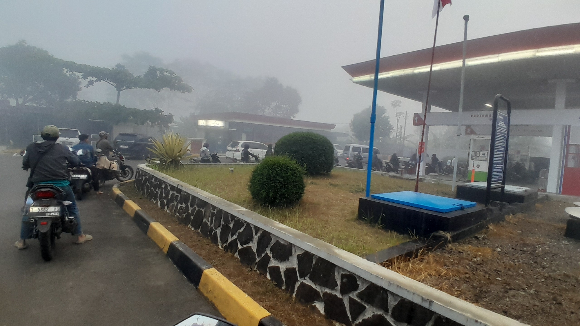 The image depicts a foggy morning scene at a Pertamina gas station in Subang, Indonesia. Several motorbikes are lined up, suggesting people are waiting to refuel or have just finished. A rider on a motorbike in the foreground is wearing a green helmet and a dark jacket, facing away from the camera. Another motorbike is positioned behind it, with a rider also facing away. In the background, a row of motorbikes and a few cars are visible near the gas station pumps. The gas station itself has a red and white canopy and is characterized by the "PERTAMINA" signage. There are also two spherical bushes planted in front of the gas station. The overall environment is hazy and muted due to the fog.