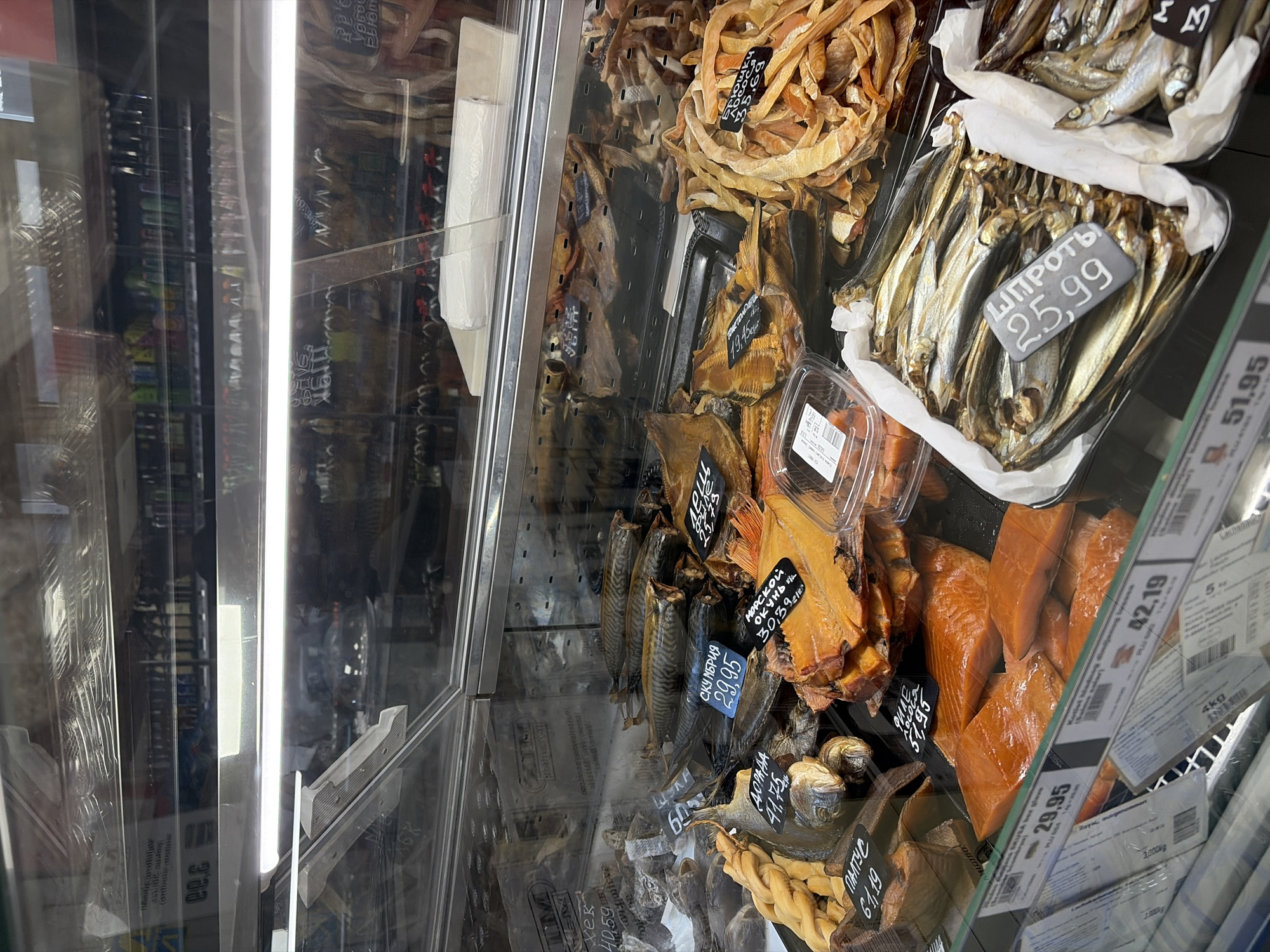 The image shows a display case filled with various types of smoked fish, presumably inside a store in Budva, Montenegro, given the Cyrillic text on the price tags. The fish are arranged in trays and include sprats, mackerel, sea bass, halibut, and salmon, with visible price tags indicating prices like 25.99 for sprats and 29.95 for mackerel, among others. The display case is well-lit by a vertical fluorescent lamp and is partially reflected on the transparent surfaces.