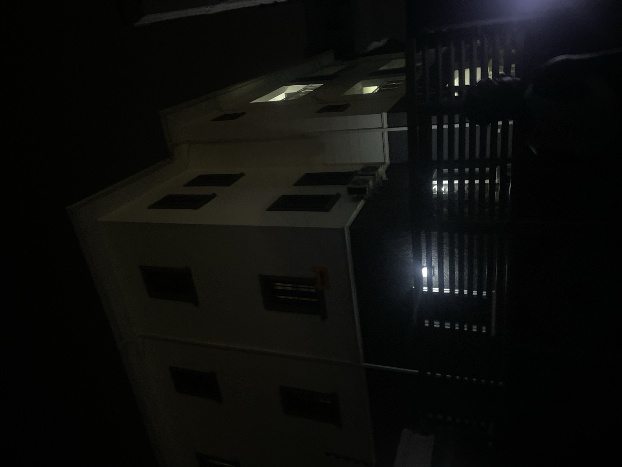 The image displays a multi-story building at night in Gwarinpa, Nigeria, enveloped in deep darkness with selective artificial illumination. The main subject is a light-colored facade of what appears to be a residential or commercial building. Several windows are visible, with some emitting bright, warm light from within, while others remain dark. Below some windows, small dark rectangular units, possibly air conditioning units or vents, are affixed to the exterior.
To the right of the building, a prominent dark structure with vertical slats, resembling a fence or railing, is visible. Behind this structure, bright light sources pierce through the slats, casting vertical streaks of light. These lights and the slatted pattern are clearly reflected on a dark, highly reflective surface in the foreground, suggesting a wet ground or a polished surface.
The scene is dominated by darkness, emphasizing the illuminated windows and the reflections, which contribute to a quiet, nocturnal ambiance. No individuals are present. The time of day is undeniably night. On one side of the building, a dark rectangular sign with faint, unreadable light-colored text can be discerned, along with a small, yellow square sticker or tag. The weather conditions are not explicitly clear, though the reflections hint at a potentially wet surface. The building's architecture is functional and common for urban settings.
