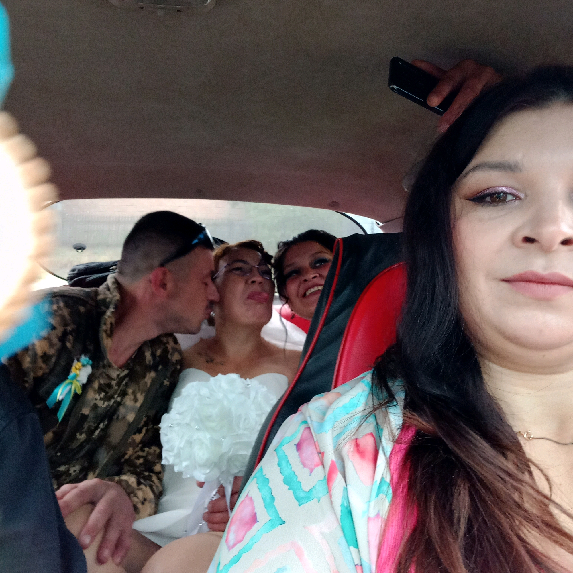 In a vehicle's interior, a man in a camouflage uniform kisses a woman wearing a white wedding dress. To their right, another woman with glasses sticks her tongue out, and to her right, a third woman smiles, holding a black smartphone. A bouquet of white flowers is held by one of the women. The individuals appear to be in Klyntsi, Ukraine.