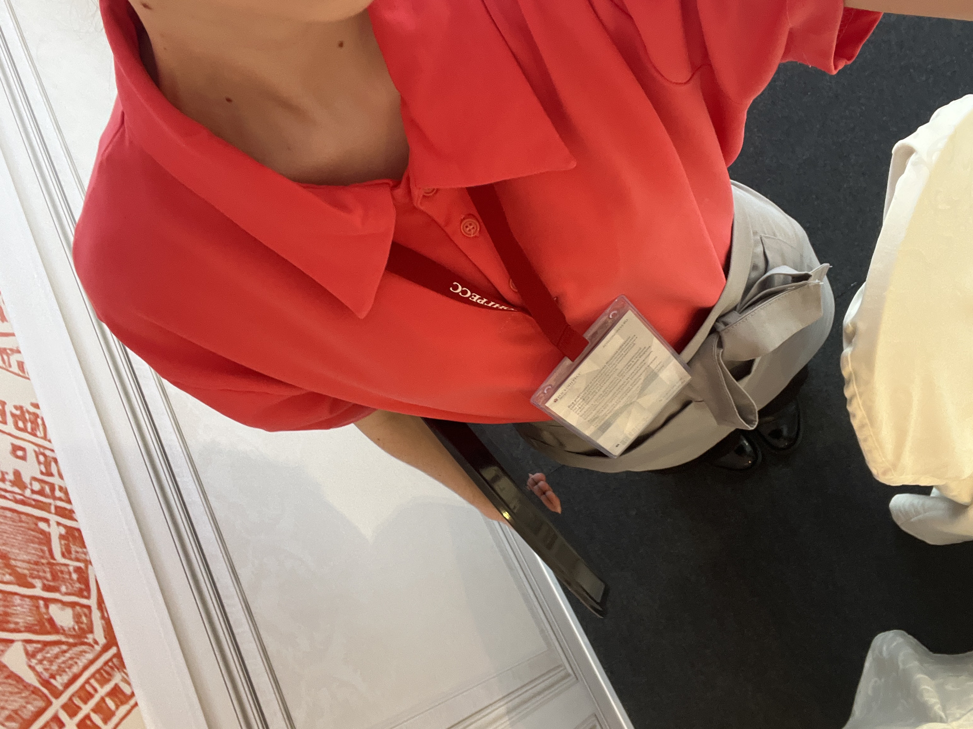 An overhead shot captures a person from the chest downwards, wearing a red short-sleeved polo shirt and light grey pants, in an indoor setting. The individual is likely at an event or work function, possibly in Shushary, Russia, given the ID badge worn around their neck.
A red lanyard with inverted white text, "HTPECC" (likely Cyrillic for an organization or event name), is visible. Attached to the lanyard is a clear plastic ID badge holder containing a white card with small, unreadable black text. The Cyrillic word "ДОКУМЕНТАЦИЯ" (Documentation) is partially visible on the badge. The person's light grey pants feature a visible pocket flap on their right side. They are holding a black tablet or similar electronic device in their left hand, down by their side. Their feet, clad in dark, polished shoes, rest on a dark grey or black carpeted floor.
To the left, a white wall features a red, intricate pattern resembling a map or architectural drawing, alongside white door frames or paneling. On the right, a white or cream-colored piece of furniture, possibly draped with a cloth, is partially visible. The scene suggests a professional or event environment.