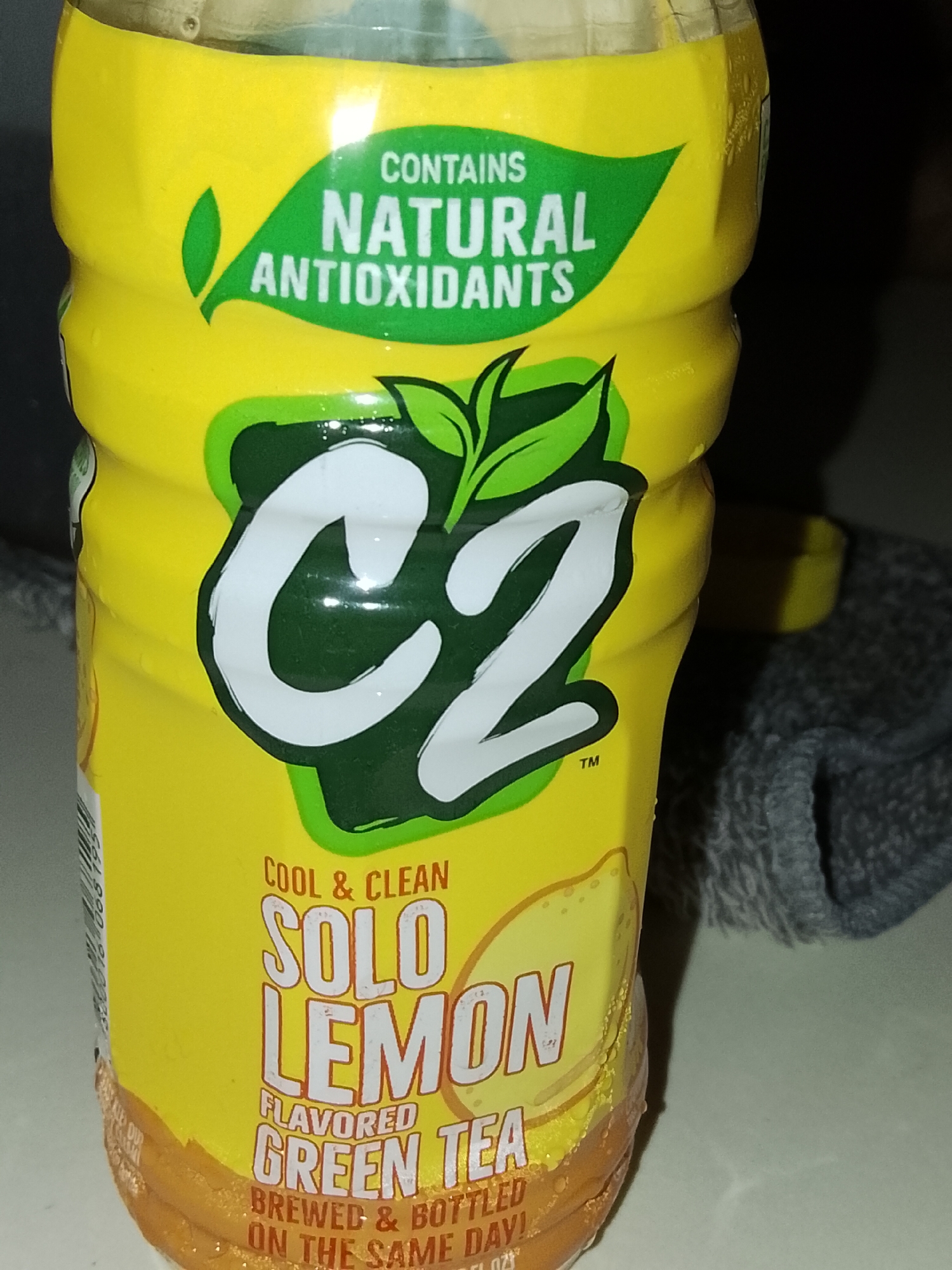 A yellow plastic bottle of C2 Solo Lemon flavored green tea is partially visible against a dark background and a gray textured object. The bottle features green leaf graphics and text indicating "CONTAINS NATURAL ANTIOXIDANTS" and "COOL & CLEAN SOLO LEMON FLAVORED GREEN TEA BREWED & BOTTLED ON THE SAME DAY!". The brand logo "C2" is prominently displayed in large white letters outlined in green. The bottle appears to be condensation-covered. The image is cropped tightly on the bottle, obscuring most of the surrounding environment. No specific location in Davao City, Philippines, can be identified from the provided image.