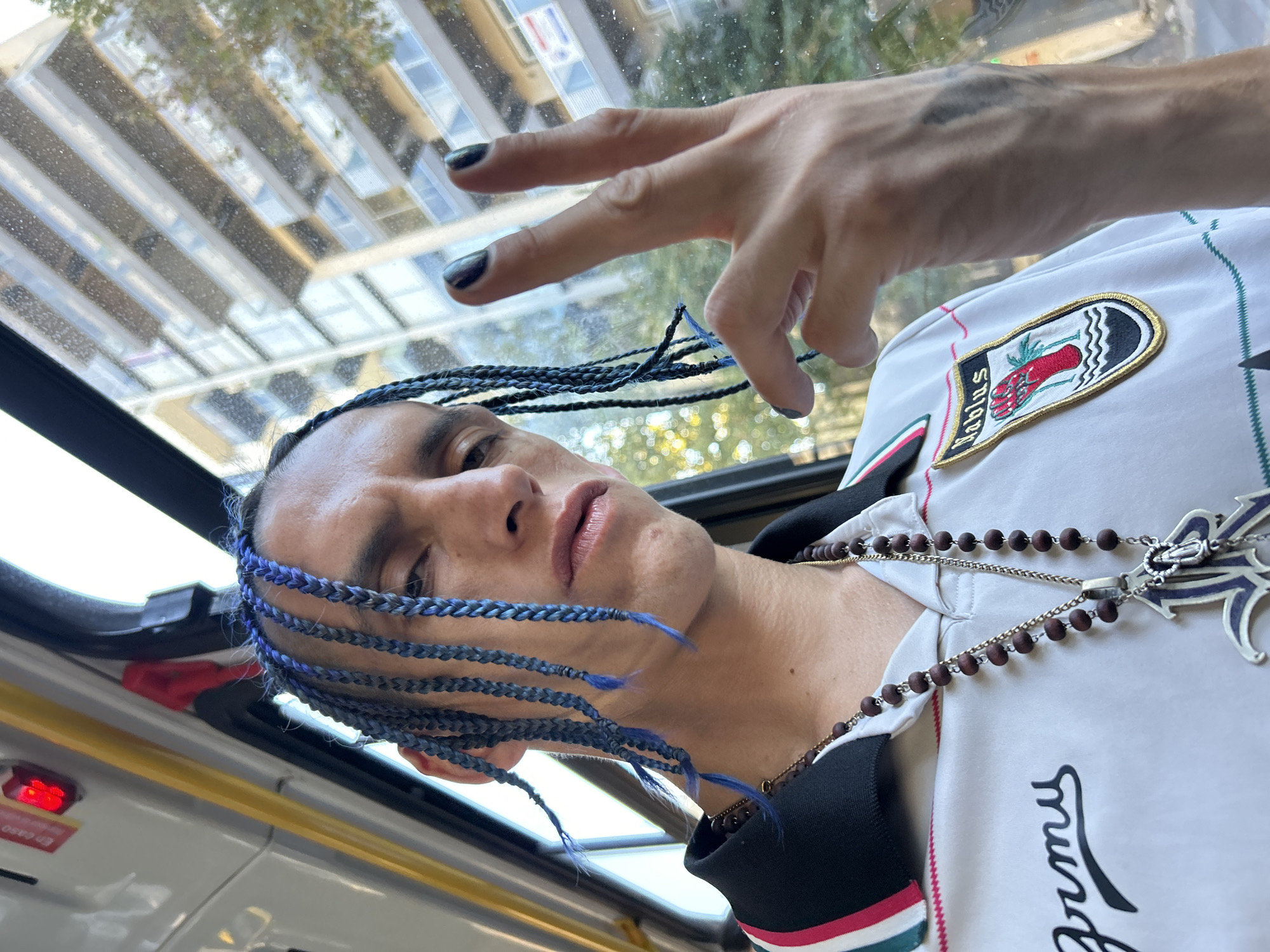 A person with blue braids and dark nail polish is photographed from a low angle in what appears to be a bus. The person wears a white shirt with a patch that reads "Fabius" and depicts a red hand. A silver necklace with wooden beads and a cross pendant hangs around the person's neck. The person's hand is raised with two fingers extended. Through the bus window, buildings and trees are visible. The bus appears to be in Buenos Aires, Argentina based on the "En caso" emergency information sticker.