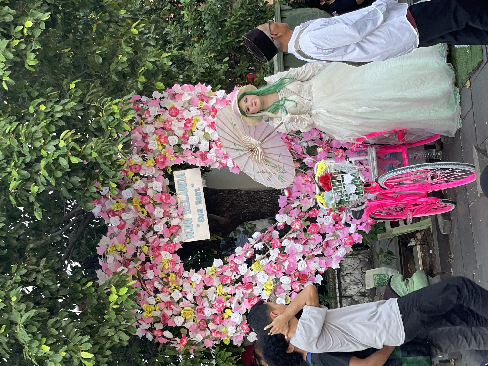 The image depicts a scene in Kota Tua, West Jakarta, Indonesia, likely during a public event or festival. In the foreground, a young woman with green-tinted hair, wearing a pale cream-colored dress and a matching hat, holds a delicate pink parasol. She is posing in front of a large heart-shaped floral arrangement composed of pink, white, and yellow artificial flowers, with a white sign reading "NONI BELANDA" and "Eva Rent" hanging from a tree trunk. To her right, an elderly man wearing a white shirt and a black peci hat is visible. In the left foreground, a man in a grey t-shirt and black pants is adjusting his hair. Behind the floral heart, a vibrant pink bicycle with a basket full of flowers is parked. The setting appears to be outdoors, with lush green trees forming a canopy overhead. The ground is paved with cobblestones, and there are various elements of urban street life present, including what appears to be a white plastic bag with green text and a metal railing. The overall atmosphere suggests a festive or commemorative occasion.