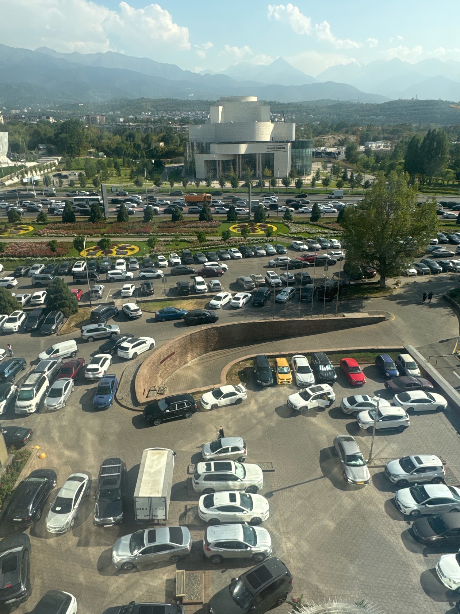 The image captures an aerial view of a large, paved outdoor parking lot in Almaty, Kazakhstan, adjacent to a modern, multi-story building complex with curved architectural elements, identified by a sign as "ALMATY THEATRE." The parking lot is filled with numerous vehicles, primarily cars, of various models and colors. Several lanes of traffic are visible, with cars moving and parked. In the background, a dense urban landscape with trees and other buildings extends towards a range of majestic, hazy mountains under a sky with scattered clouds. The overall scene depicts a busy urban environment with a prominent cultural landmark and a backdrop of natural scenery.