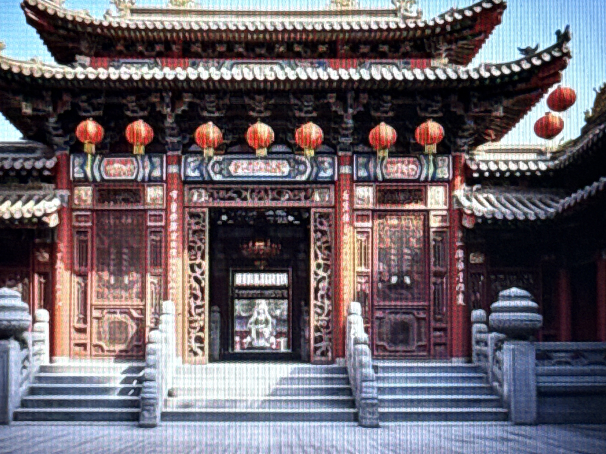 A large, ornate, traditional Chinese temple complex is shown. The architecture features multiple tiers of layered, curved roofs, elaborately decorated with white trim and upturned eaves. Red pillars and wooden beams support the structure, and numerous red lanterns are hung from the eaves, some casting a warm glow. Intricate carvings and decorative panels adorn the facade of the main entrance, which is framed by large, red wooden doors. A set of wide, stone stairs leads up to the entrance, flanked by stone balustrades with decorative finials. Within the open archway of the entrance, a statue of a seated figure is visible, illuminated from within. The ground in front of the temple is paved with a mosaic of small, grey stones. The sky is a clear, bright blue.