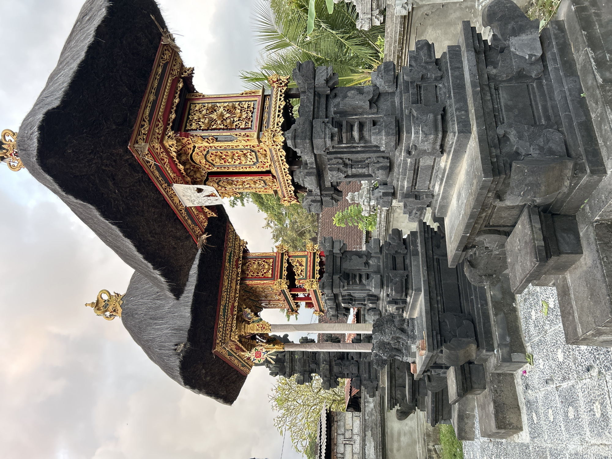The image shows a detail of two ornate Balinese shrines with thatched roofs and gold-leaf decorations. The shrines are made of dark stone and are situated on a cobblestone patio. In the background, there are palm trees and a cloudy sky. The shrines are located in Abiansemal, Indonesia, a region known for its traditional Balinese architecture and temples.