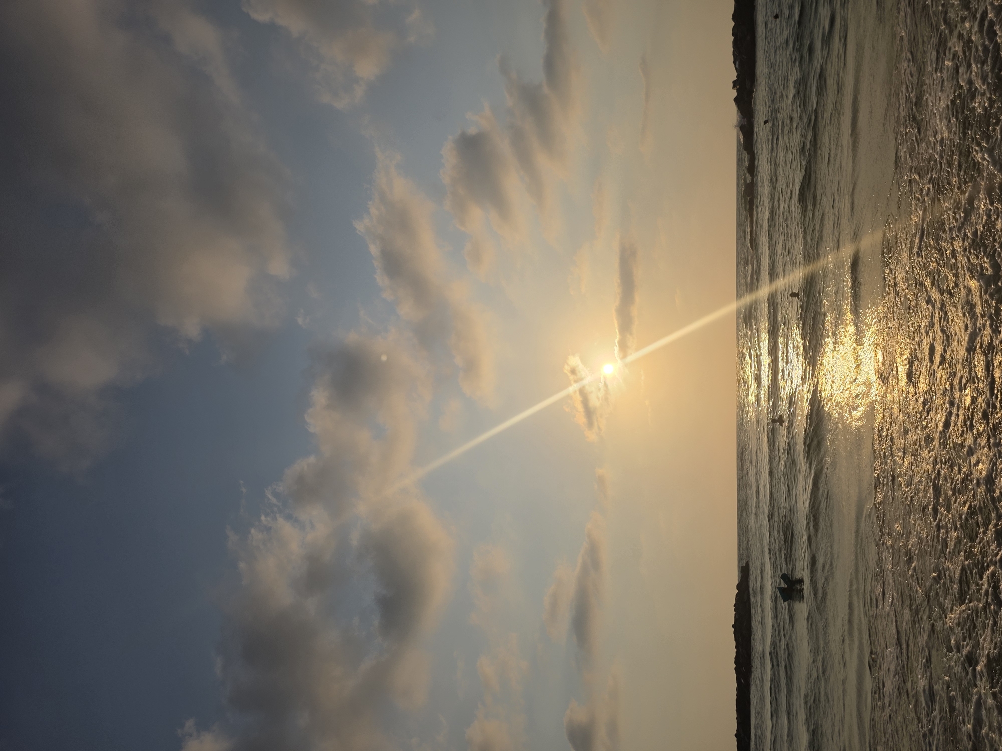 A vertical, eye-level, medium shot captures a sunset over the Mediterranean Sea at a beach in Ashkelon, Israel. The sun is low on the horizon, casting a golden glow on the water, with a bright ray of light extending upwards into the sky. The sky is a gradient of blue and pale orange, with scattered, fluffy clouds tinged with yellow and gray.

The sea is moderately choppy, with waves breaking on the shore, creating foamy whitecaps. In the distance, dark, rocky jetties jut out into the water, breaking the waves. Several individuals are visible in the water, appearing to be swimming or engaging in water activities. One person is on a blue flotation device, and at least three other figures are partially submerged, with only their heads visible. The foreground of the image shows the wet sand of the beach, with foam from the breaking waves. The overall atmosphere is tranquil and serene, depicting the end of a day at the beach.