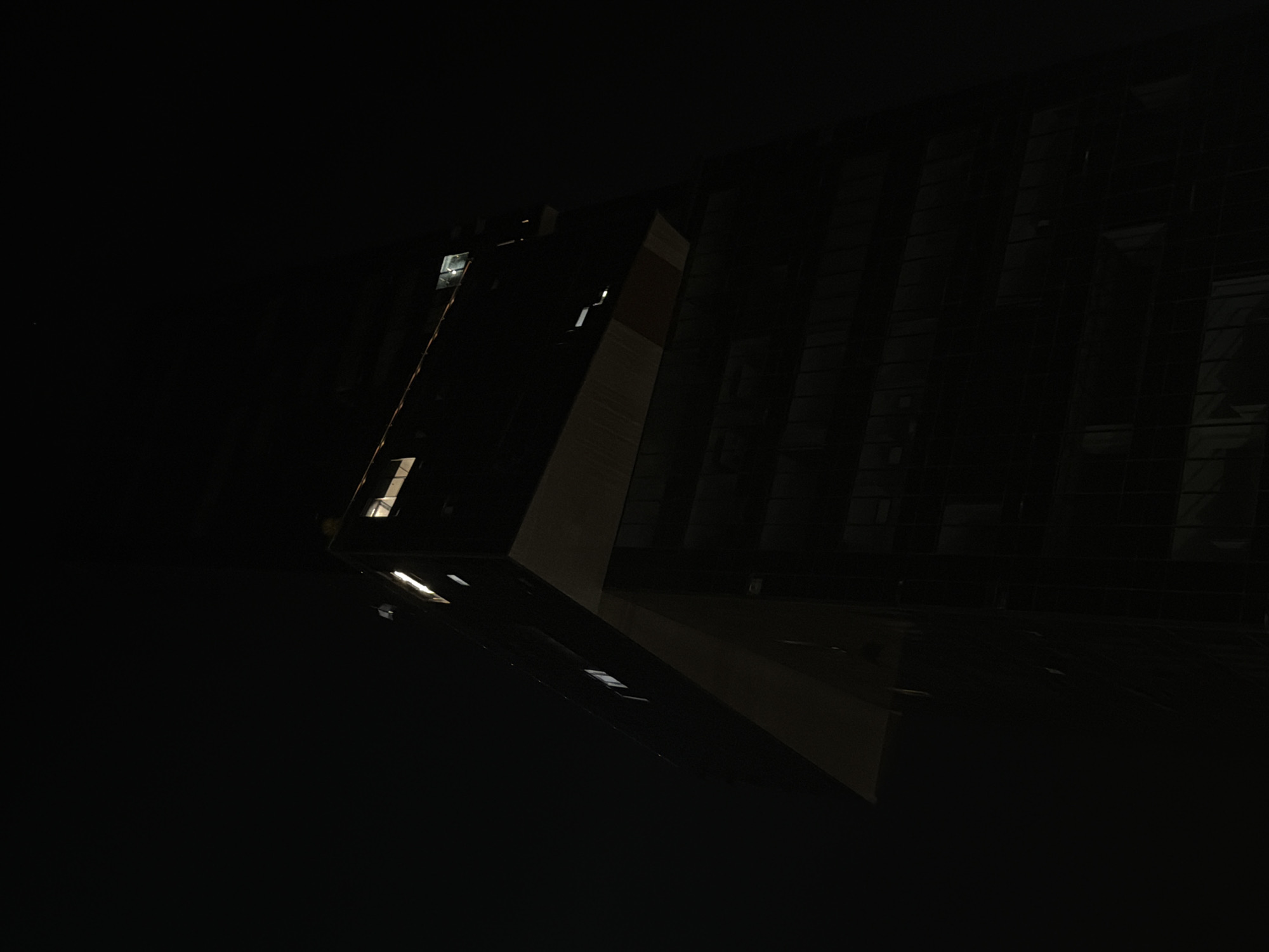 The media file presents a dark, nocturnal view of a modern building in Puebla, Mexico. The scene is dominated by deep shadows, with minimal artificial light sources illuminating aspects of the architecture.
The primary subject is a contemporary building featuring sharp, geometric lines and reflective surfaces, likely glass or polished dark stone. Its structure appears to have angled sections, creating a dynamic, almost abstract form. Several rectangular windows or internal light fixtures are visible, emitting bright white and yellowish light against the prevailing darkness. One prominent architectural element is a lighter-toned, diagonal section of the building, possibly made of concrete or a contrasting paneling material, which breaks the dark monotony.
In the lower portion of the image, a highly reflective, dark surface is visible, possibly wet ground, a polished floor, or a body of water. This surface clearly reflects the building's lights, creating elongated, distorted streaks of illumination that add to the abstract quality of the composition.
The time of day is clearly night. No people or specific activities are depicted; the scene is static and quiet. There are no visible weather cues other than the reflective surface which could suggest dampness or a highly polished material. No visible text is present in the image. The overall impression is one of modern architecture highlighted by artificial light in an urban night setting.