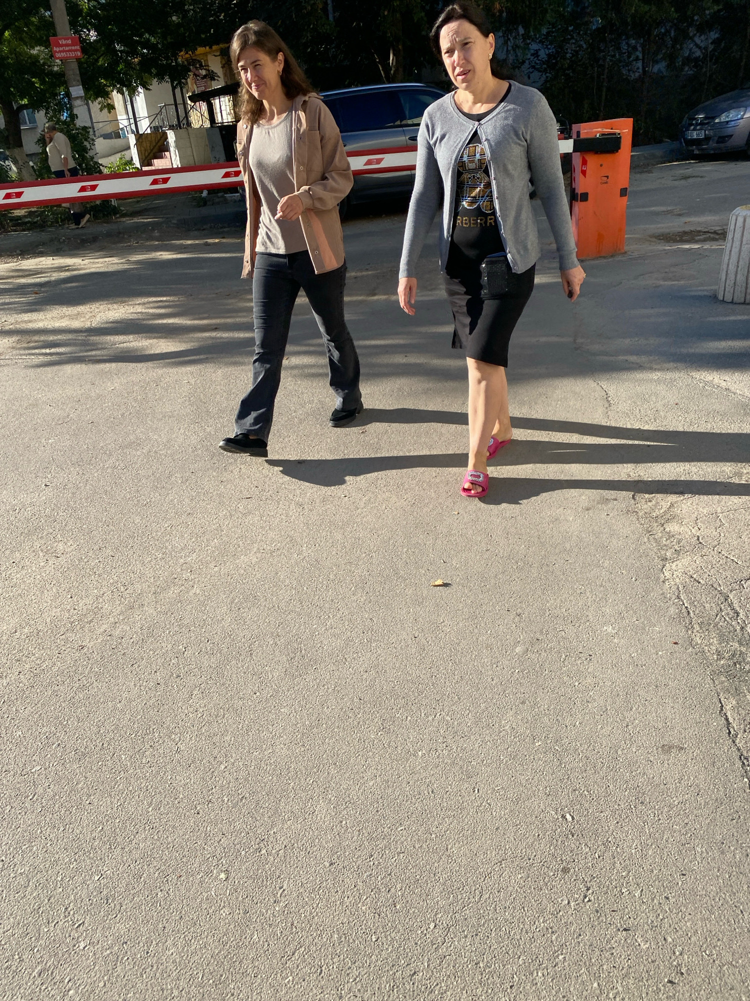 Two women walk on an asphalt surface, possibly a sidewalk or road, in Chișinău, Moldova. The woman on the left, with brown hair, wears a tan jacket over a gray t-shirt and dark pants. The woman on the right, with dark hair, wears a gray cardigan over a black t-shirt and a black skirt. She also has a black pouch attached to her waist and is wearing bright pink sandals. In the background, there is a red and white barrier gate, a silver car, trees, and a building. The scene appears to be outdoors during the daytime, with sunlight casting shadows on the ground.