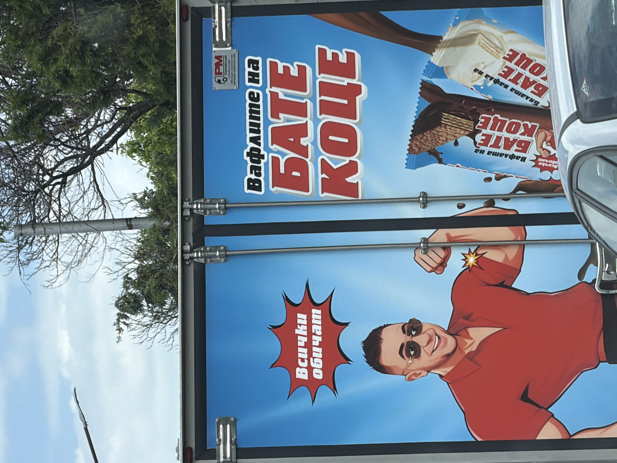 The image shows a close-up of a vehicle's back door, likely a delivery truck, adorned with an advertisement for "Bate Koce" wafers. The advertisement features images of the chocolate and vanilla wafers, the phrase "Вафлите на БАТЕ КОЦЕ" (Wafers of Bate Koce), and a cartoon image of a muscular man wearing sunglasses, flexing his arm, and smiling. A speech bubble says, "Всички обичат" (Everyone loves). The vehicle is parked outdoors with a partial view of a tree and sky. The image was likely taken in Plovdiv, Bulgaria, given the language and the name of the product.
