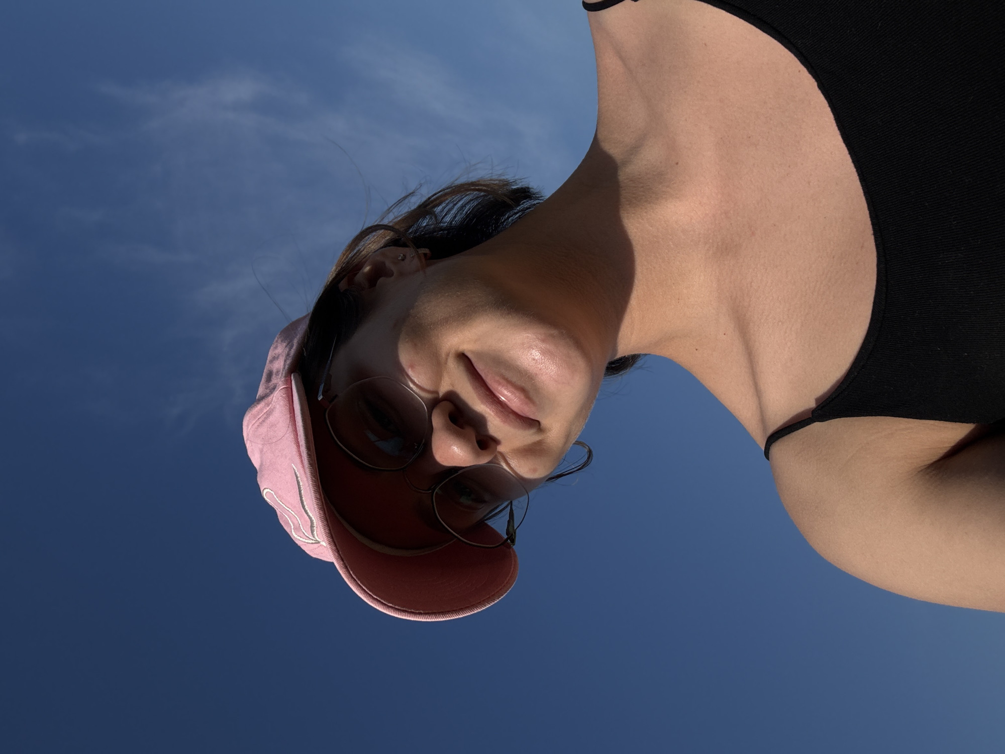 An upward-angle, medium shot captures a young woman wearing a pink baseball cap and round-rimmed sunglasses, looking down at the camera with a slight smile. She is wearing a black tank top. Her hair is brown and appears windswept. The background is a clear blue sky with a few wispy clouds. The image does not provide any visual information to suggest a location in the city of Докша, Russia.