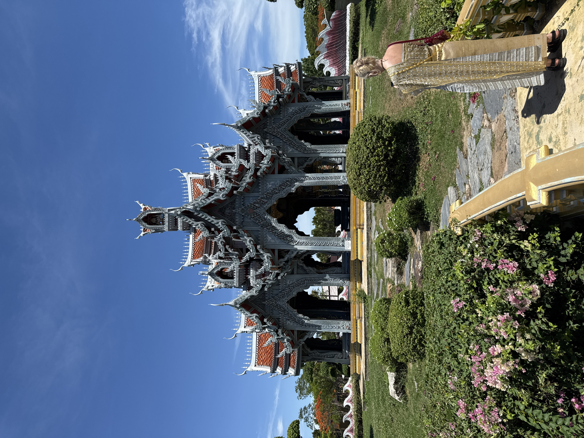 A woman in traditional Thai attire stands next to a highly ornate pavilion. The pavilion features multiple layers of intricate grey architectural details and red tiled roofs under a clear blue sky with some wispy clouds. The surrounding environment consists of meticulously landscaped greenery, including shaped shrubs and flowering plants. The setting suggests a cultural or historical site within the Bang Pu Subdistrict Municipality, Thailand.