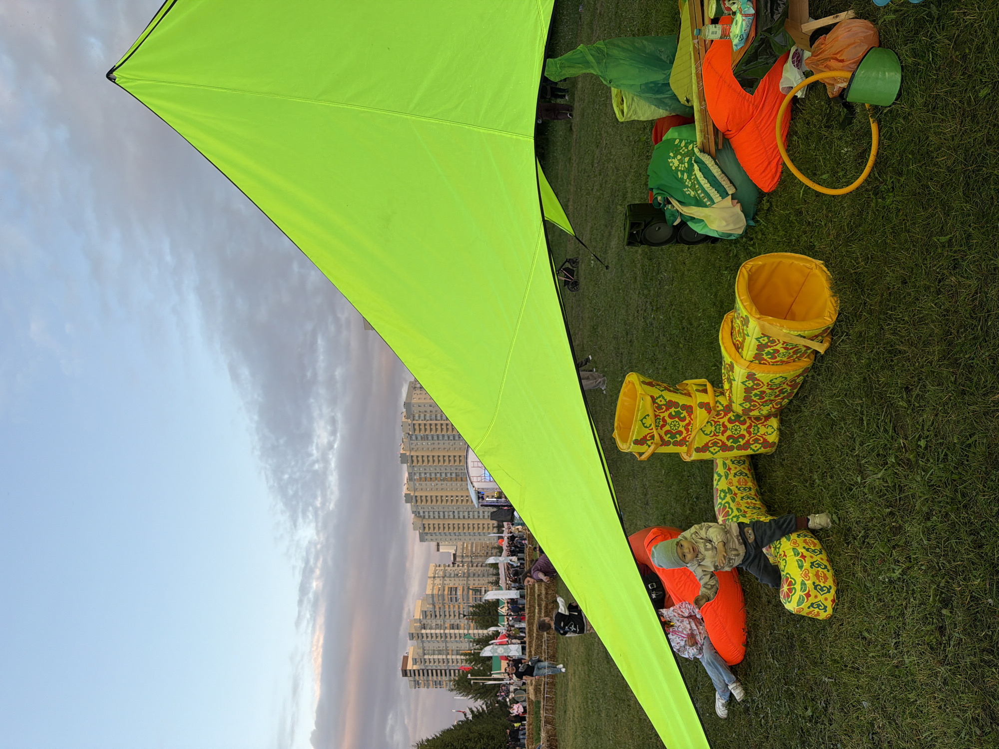 An outdoor event is taking place on a grassy field with a large, bright green tent canopy in the foreground. Several buildings in the background suggest an urban setting. On the grass, a child in a patterned hoodie and grey hat is playing near a yellow, patterned play tunnel. Two adults are sitting on an orange beanbag chair to the left. To the right, there are various items scattered on the grass, including bags, a speaker, and a green rain poncho. A crowd and stage area are visible further in the background, indicating a public gathering or festival. The sky is cloudy with hints of sunset colors.