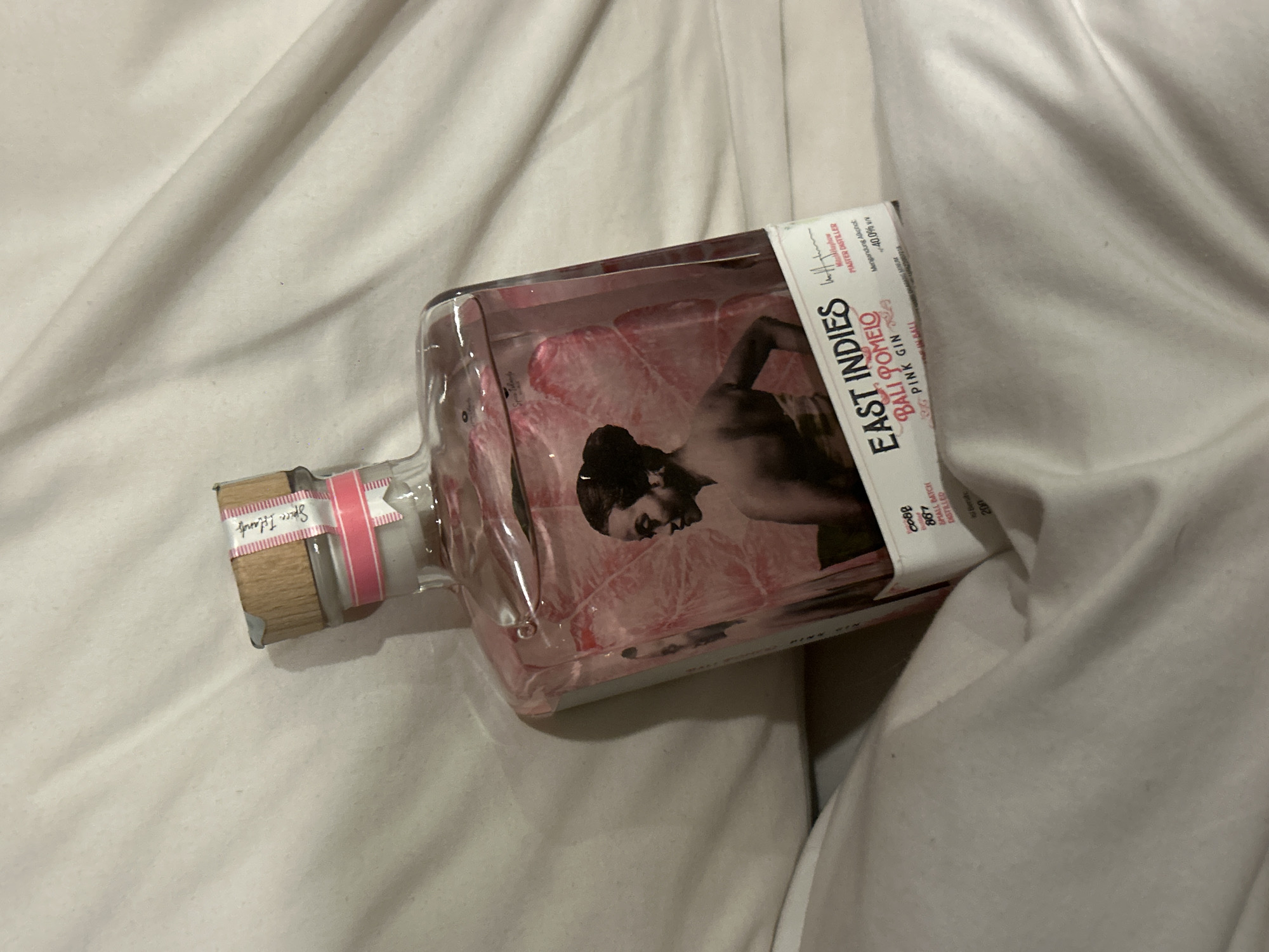 The image shows a partially visible, prone, rectangular clear glass bottle of East Indies Bali Pomelo Pink Gin on a white, creased fabric surface, likely a bed sheet. The bottle's label features a black-and-white image of a person, the text "EAST INDIES BALI POMELO PINK GIN," and smaller text mentioning "MASTER DISTILLER," and "40.0%." A "Spice Islands" label is near the bottleneck, decorated with a pink ribbon. A small label on the bottle mentions that it is from a small batch distilled in 200 with the bottle number being 006887, possibly produced in North Kuta, Indonesia.