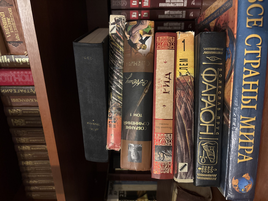 The image displays a static arrangement of books within an indoor environment, specifically identified as Velikiye Luki, Russia. In the foreground, seven books are stacked horizontally, spines facing the viewer. The topmost book has a black hardcover with "Л. ТЕРНИК" (L. TERNIK) visible. Below it is an orange-red book with patterned artwork. The third book is brown, titled "СОБРАНИЕ СОЧИНЕНИЙ ТОМ 1" (COLLECTED WORKS VOLUME 1), featuring an illustration of a quill pen and two figures embracing. The fourth book is red, labeled "МАЙН РИД" (MAYNE REID) with "Всадник без головы" (The Headless Horseman) also visible. The fifth book, with a multi-toned spine, shows "С. ЛЕМ" (S. LEM). The sixth book is black with prominent gold lettering spelling "ФАРАОН" (PHARAOH) and Egyptian-style symbols. The bottom book in the stack is blue, titled "ЗАГАДКИ СТРАН МИРА" (MYSTERIES OF THE COUNTRIES OF THE WORLD) and includes a globe illustration.
In the background, a wooden bookshelf is visible, housing numerous other books arranged vertically. Titles discernible on the upper shelf include "Лондон" (London), "М. Карамзин" (M. Karamzin), "Трагедии" (Tragedies), and "А. Пушкин" (A. Pushkin). To the right, additional books with red and white spines are vertically oriented, some bearing the word "НОВАЯ" (NEW). No people are present, and no active interactions are depicted. The scene is illuminated by ambient light.