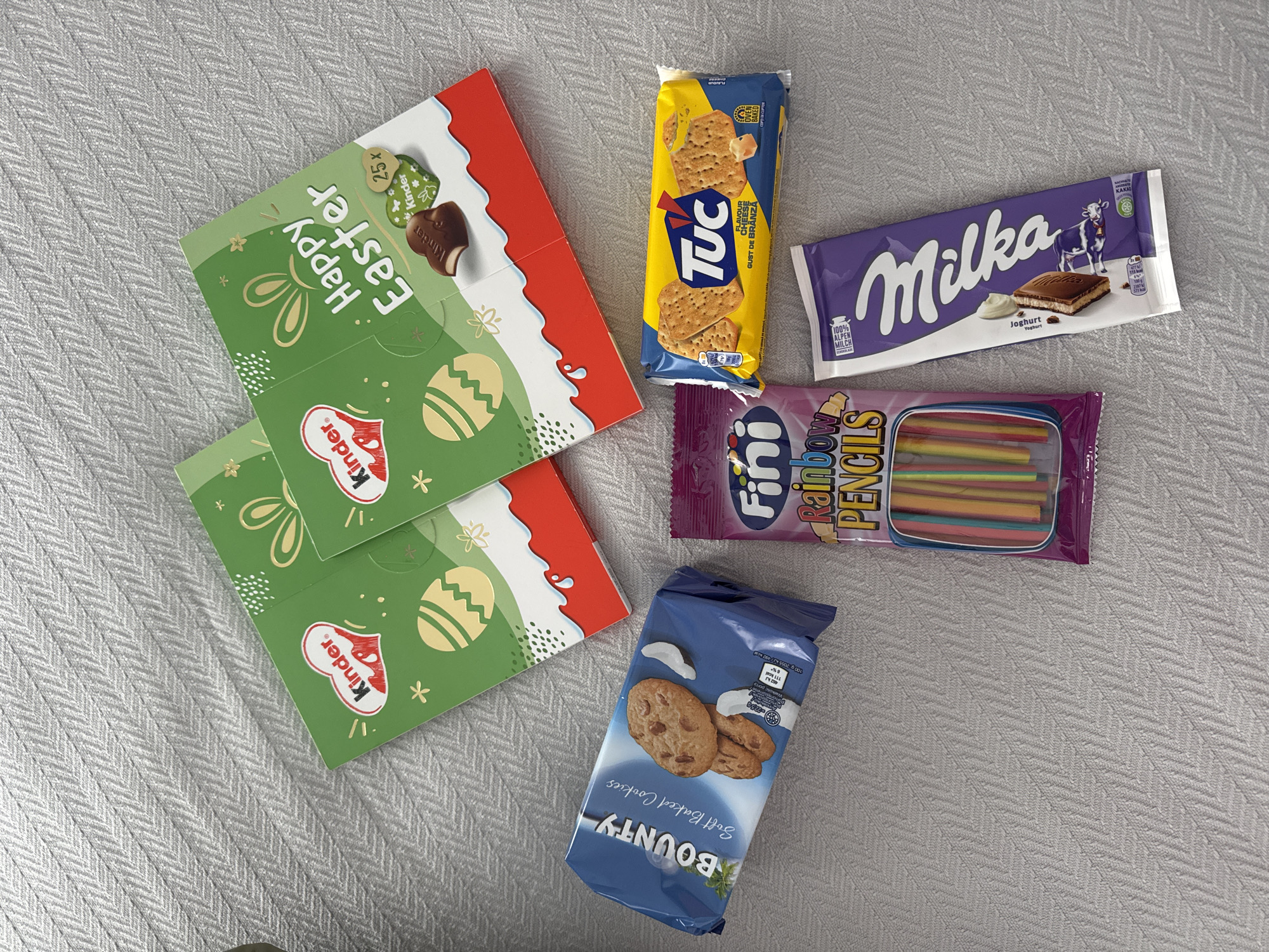 The image shows an overhead view of a collection of snacks and treats arranged on a textured, light-colored surface, possibly a fabric or tablecloth. The items include two boxes of Kinder chocolate Easter treats with green packaging and the words "Happy Easter" on them, a packet of "TUC" cheese-flavored crackers, a "Milka" yogurt chocolate bar, a bag of "Fini Rainbow Pencils" candy, and a bag of "Bounty Soft Baked Cookies."
There is no indication of the image being located in Blagoevgrad, Bulgaria.